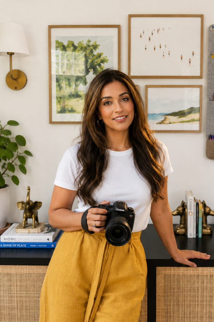 A woman with long brown hair holding a camera in a room with art and books on the wall behind her.