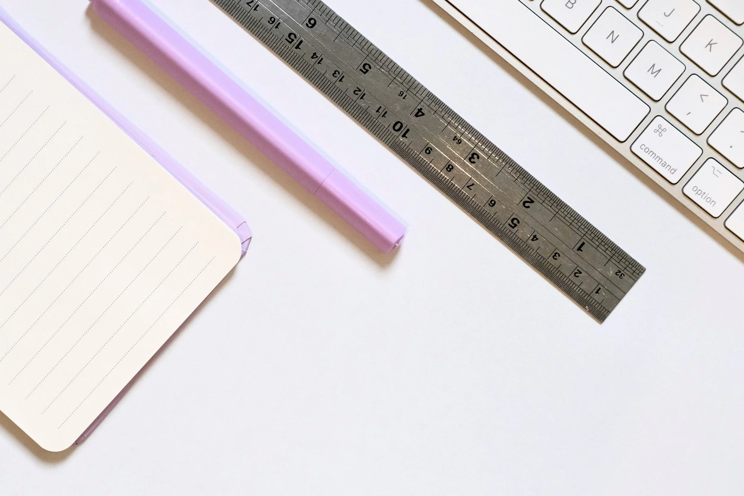 A silver metal ruler, a purple pen, a white computer keyboard, and a lined notebook are arranged on a white surface.