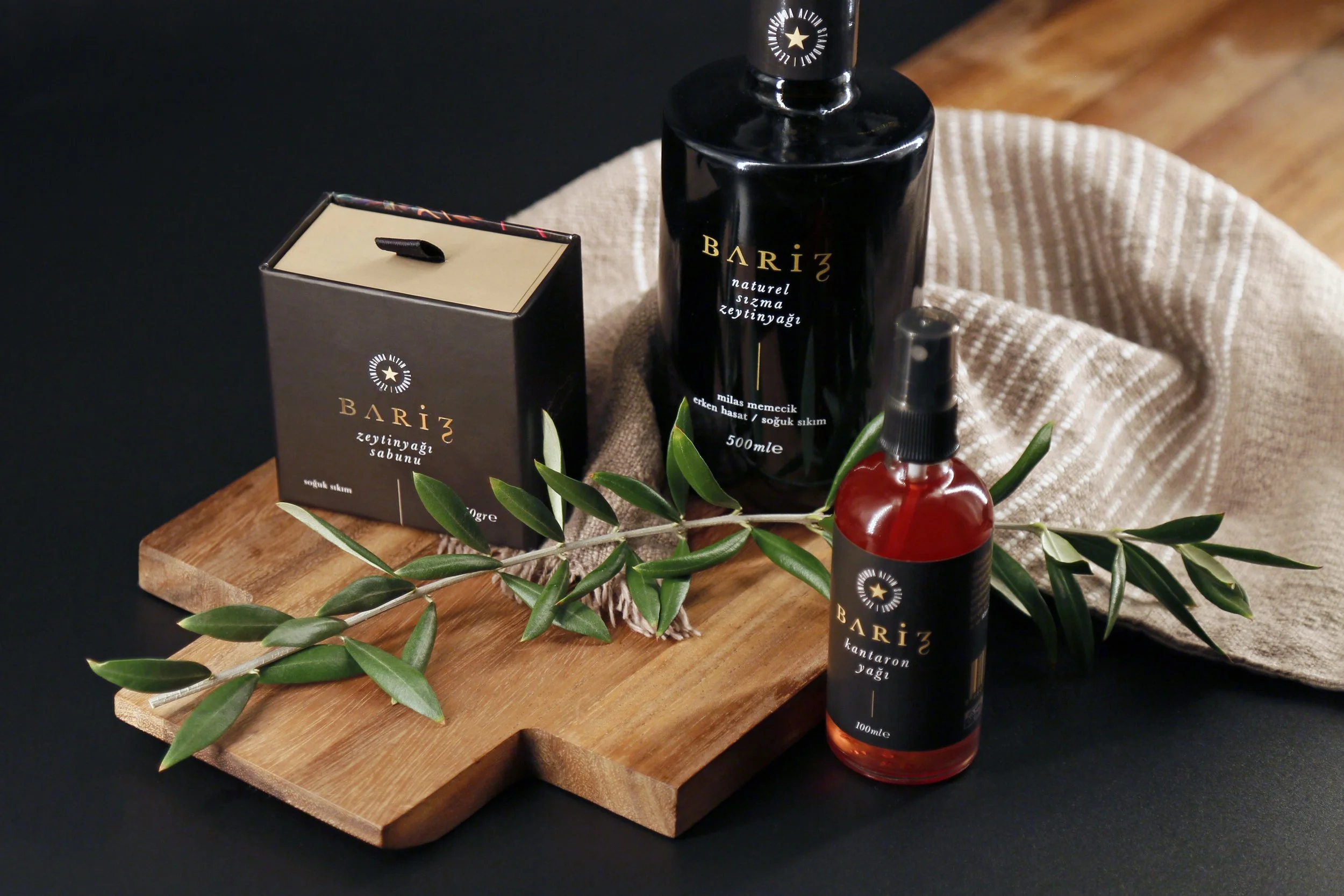 Black Barij soap box, black bottle of Barij olive oil, red bottle of Barij oil, wooden board, olive branch, and cloth on black and wooden surface.