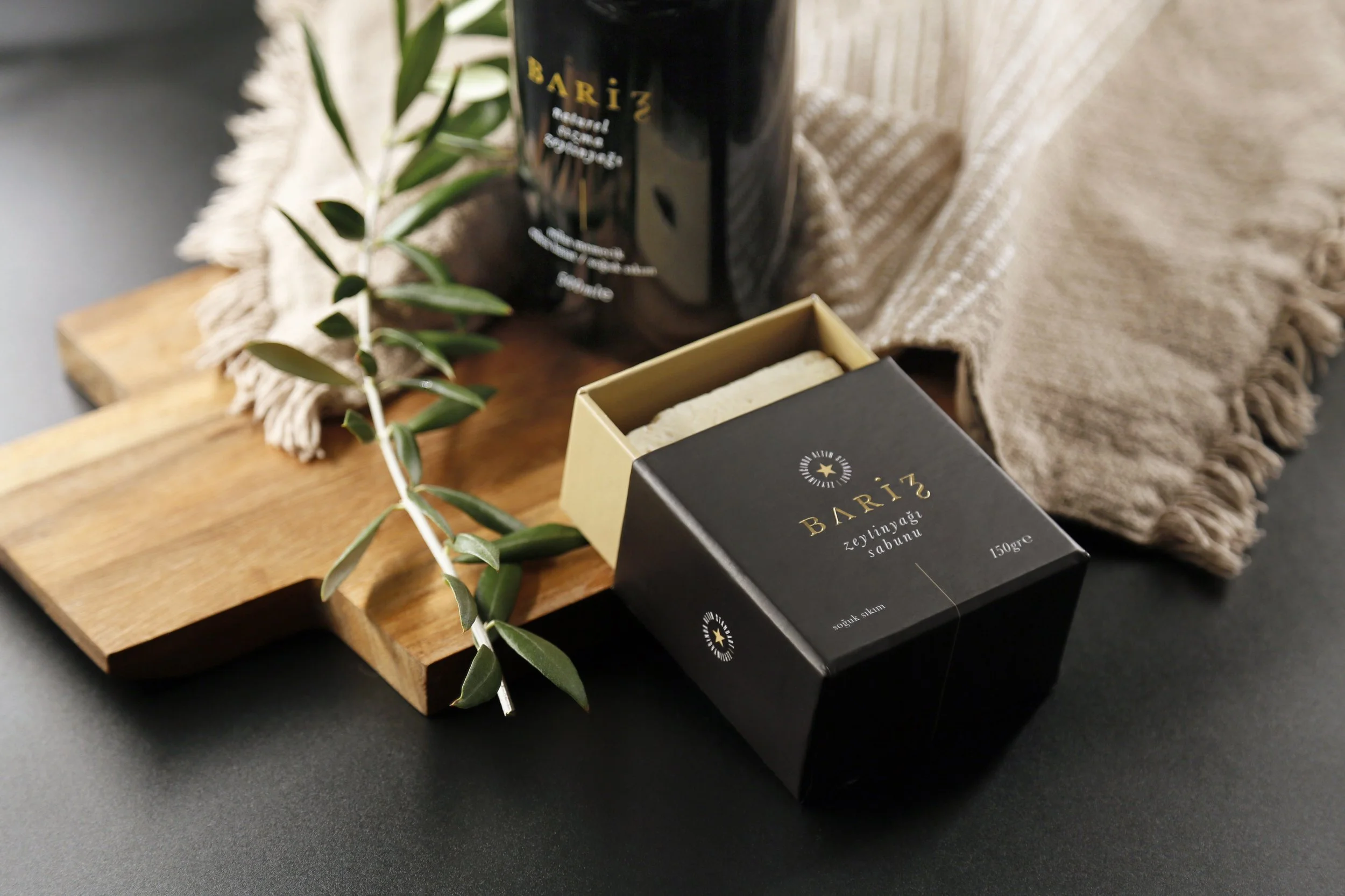 Bariz olive oil soap and black bottle on a wooden board with a beige fringed cloth and an olive branch, set on a black surface.