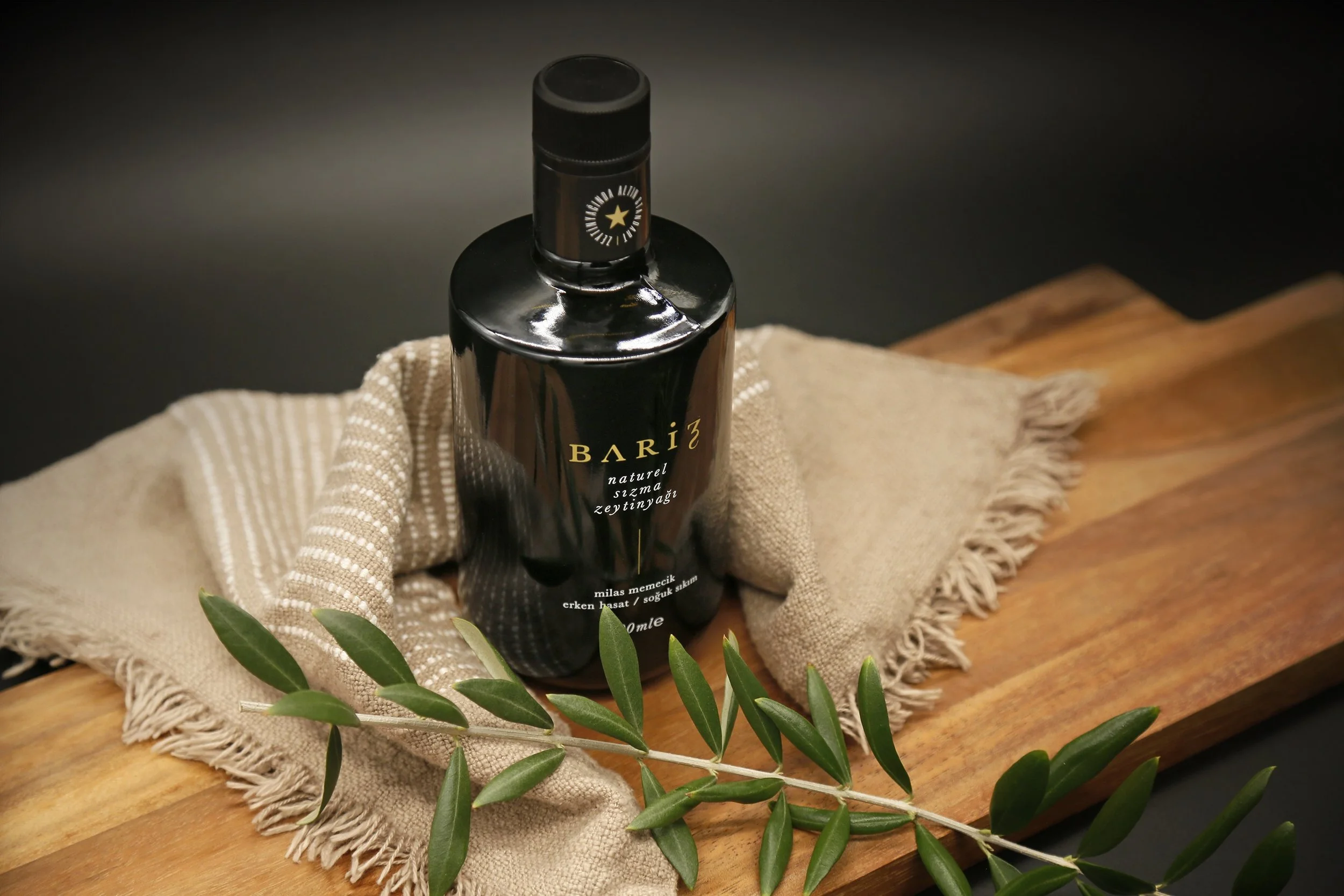 Black bottle of Bariz olive oil on a wooden board, with a beige cloth and olive branch.
