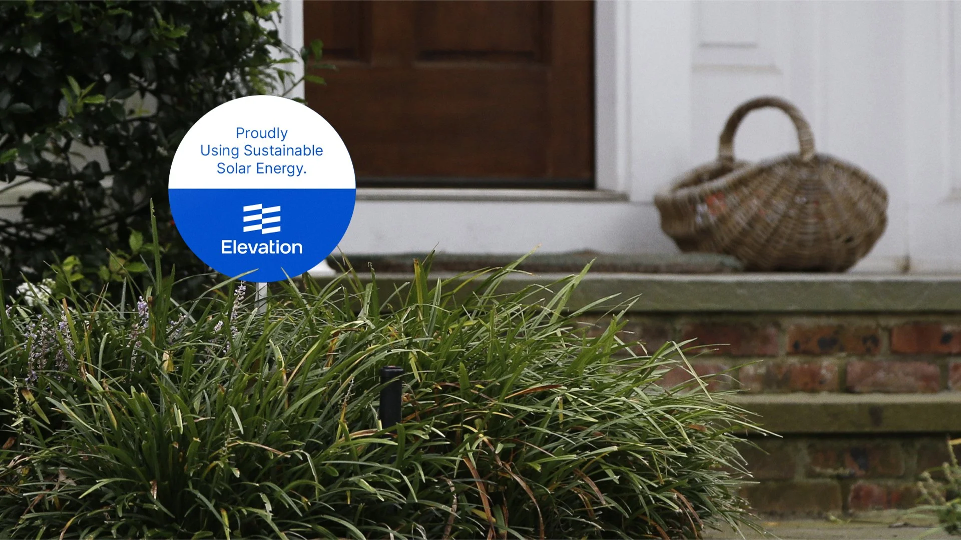 Sign in garden reading 'Proudly Using Sustainable Solar Energy. Elevation' with a wicker basket on steps in background.