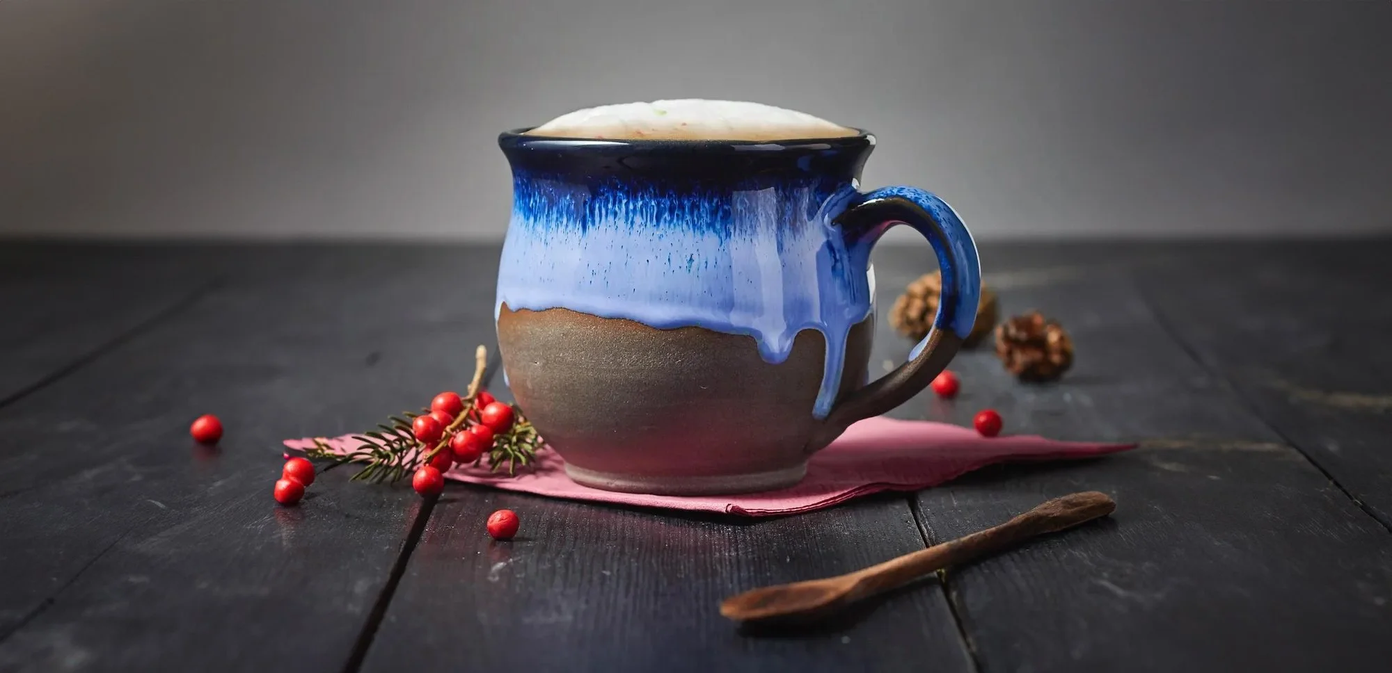 Ceramic mug with blue glaze on a wooden table with red berries, a pink napkin, and a wooden spoon.