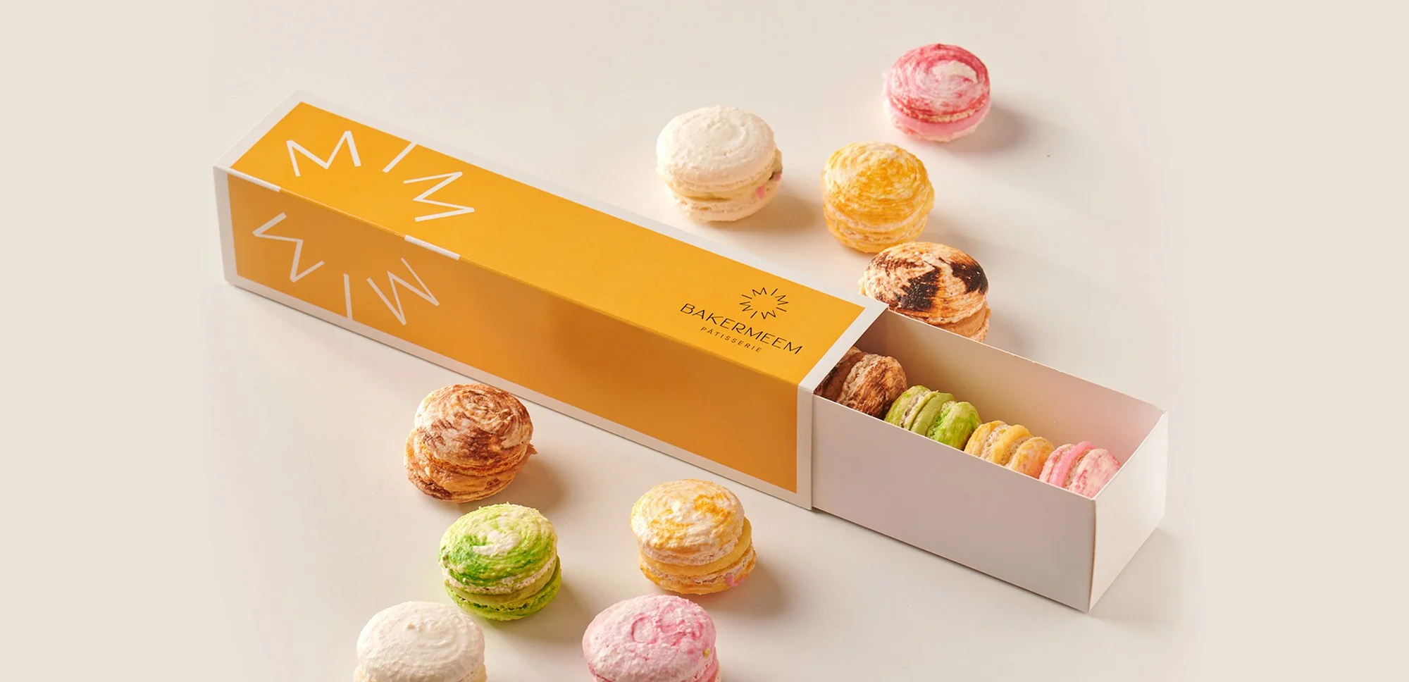 Assorted macarons in a yellow and white box with "BAKERMEEM PATISSERIE" branding, surrounded by loose macarons on a beige background.