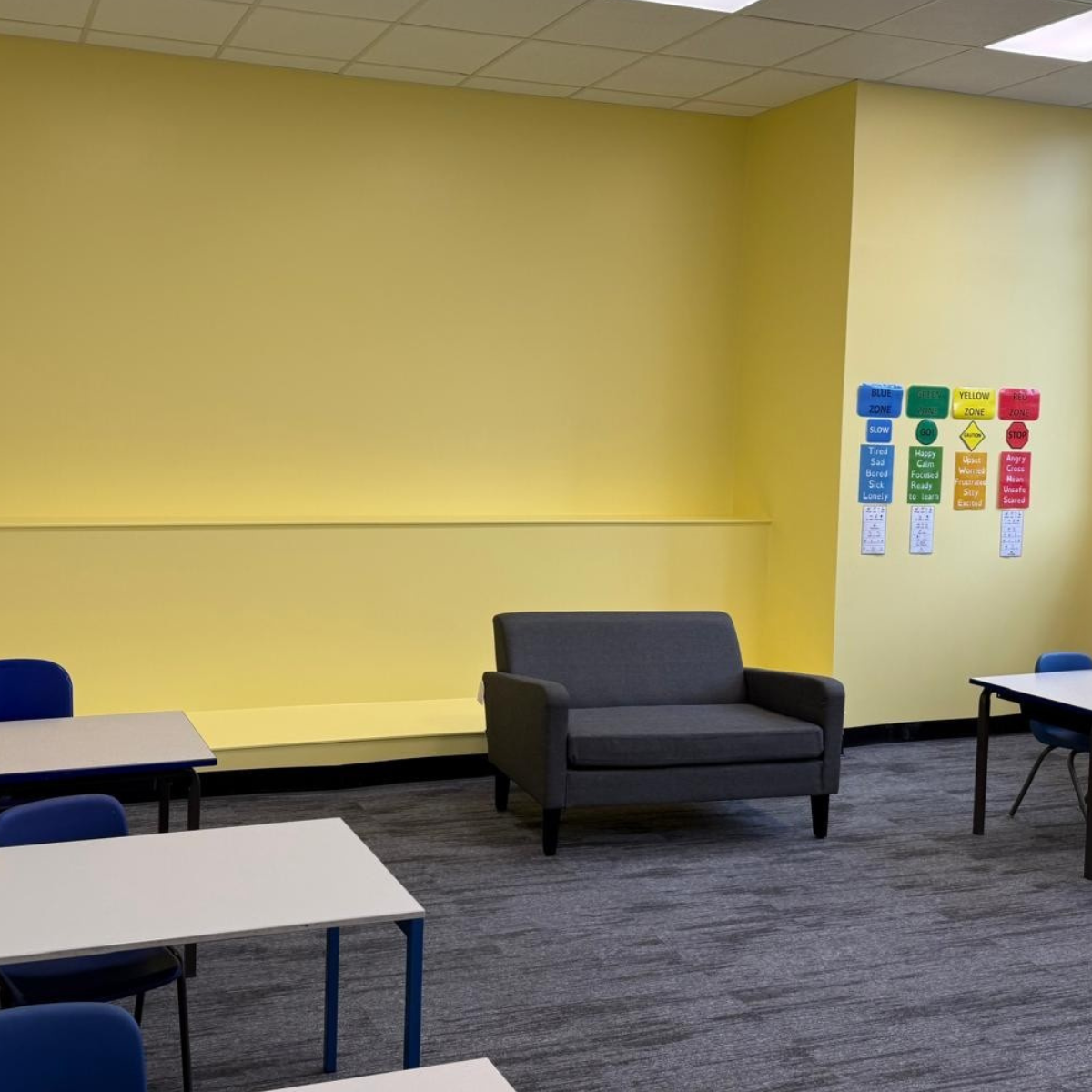 A classroom, with 4 desks, 4 chairs, a sofa and yellow walls.