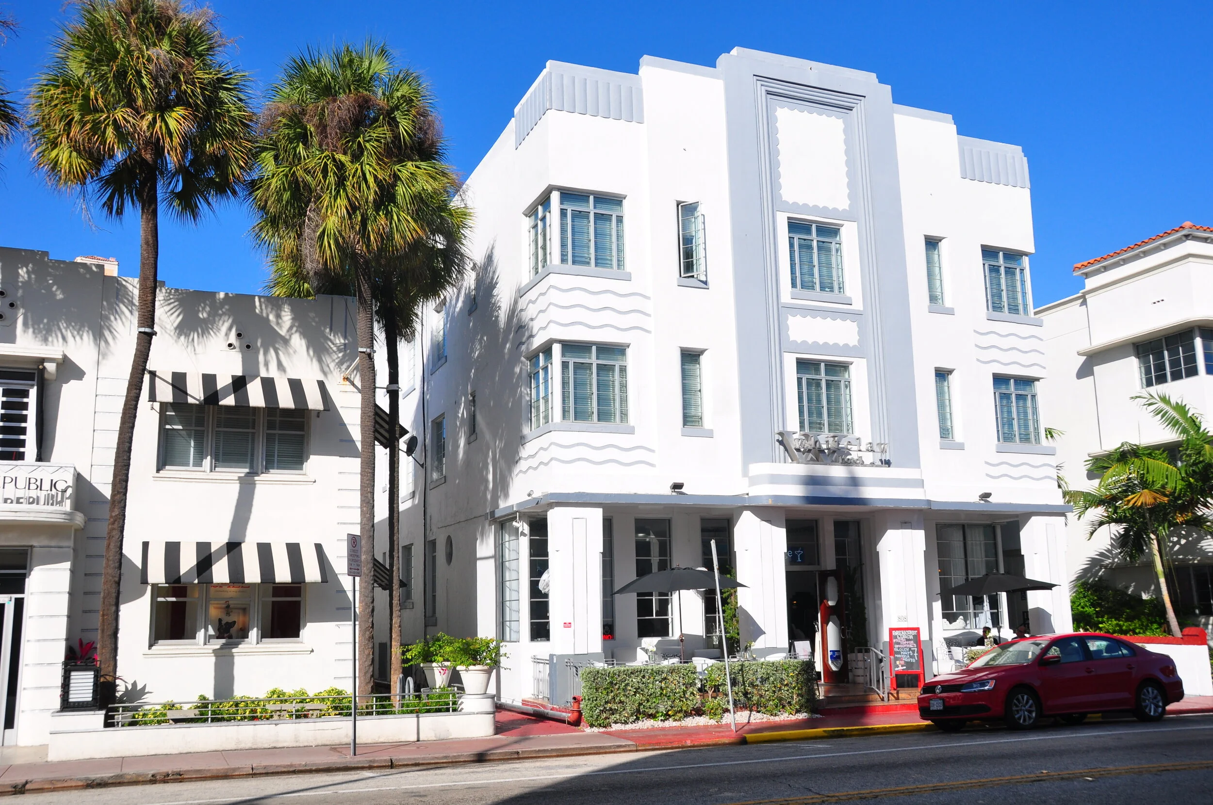 White art deco style building with large windows and exterior decor, palm trees in front, outdoor seating with umbrellas, a red car parked on the street, and a bright blue sky.