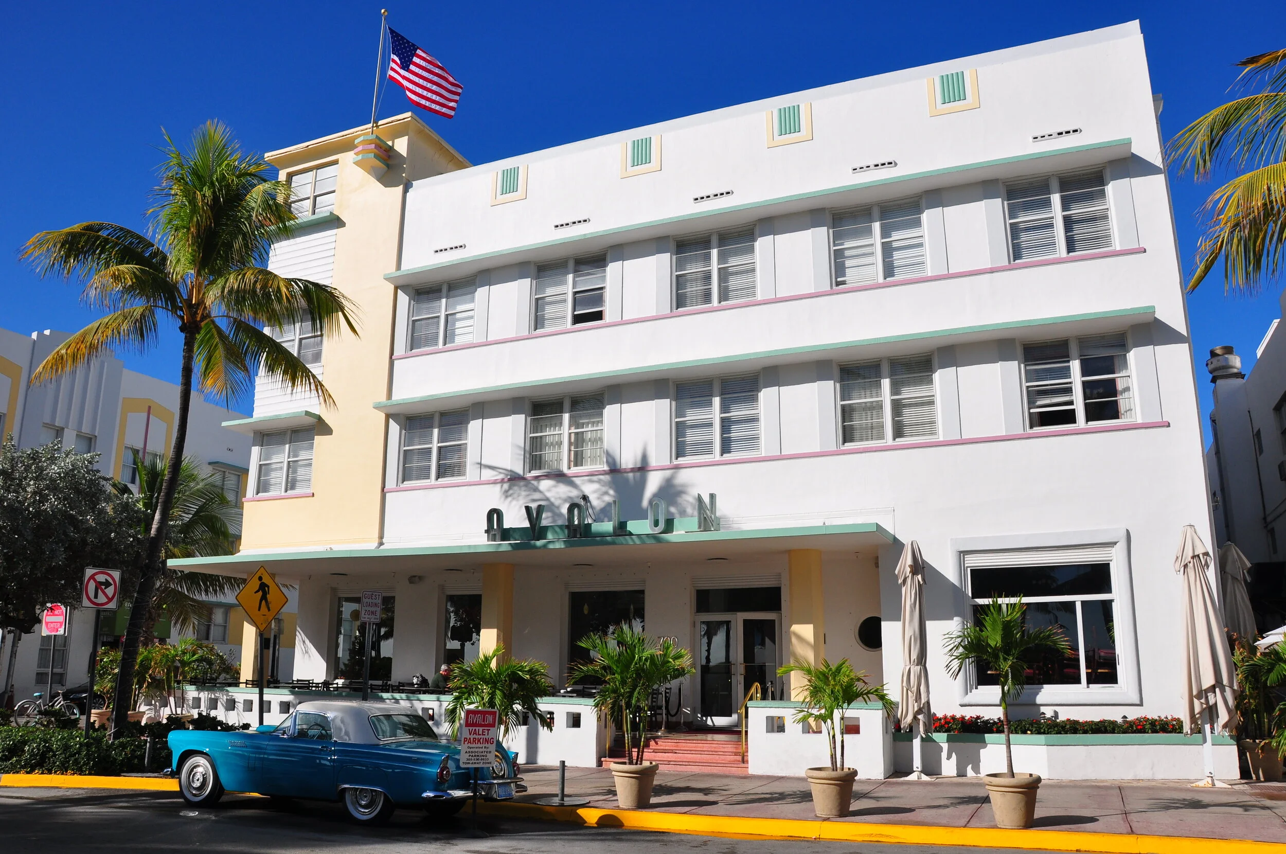 White Art Deco-style building with green accents and a sign that reads 'Lyton' at the entrance, tropical palm trees, a vintage blue car parked in front, and an American flag on the rooftop, under a bright blue sky.