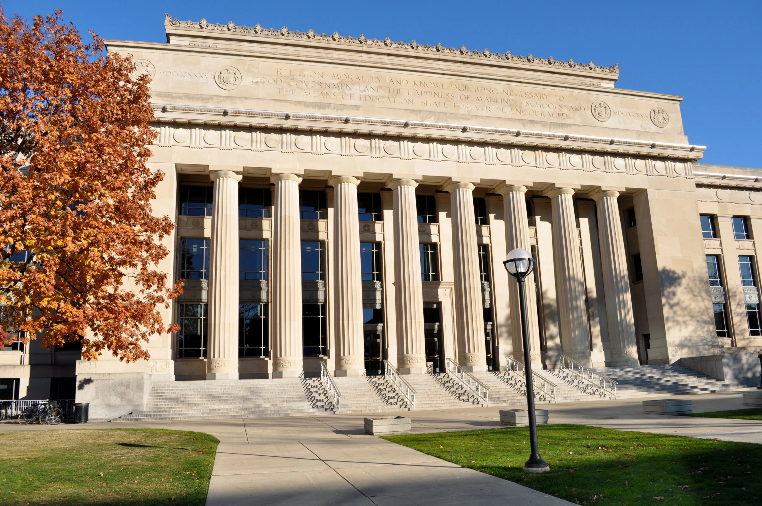 Front view of a neoclassical building with tall columns, stairs, and engraved reliefs, possibly a government or institutional building, with trees displaying fall foliage in the foreground and clear blue sky overhead.