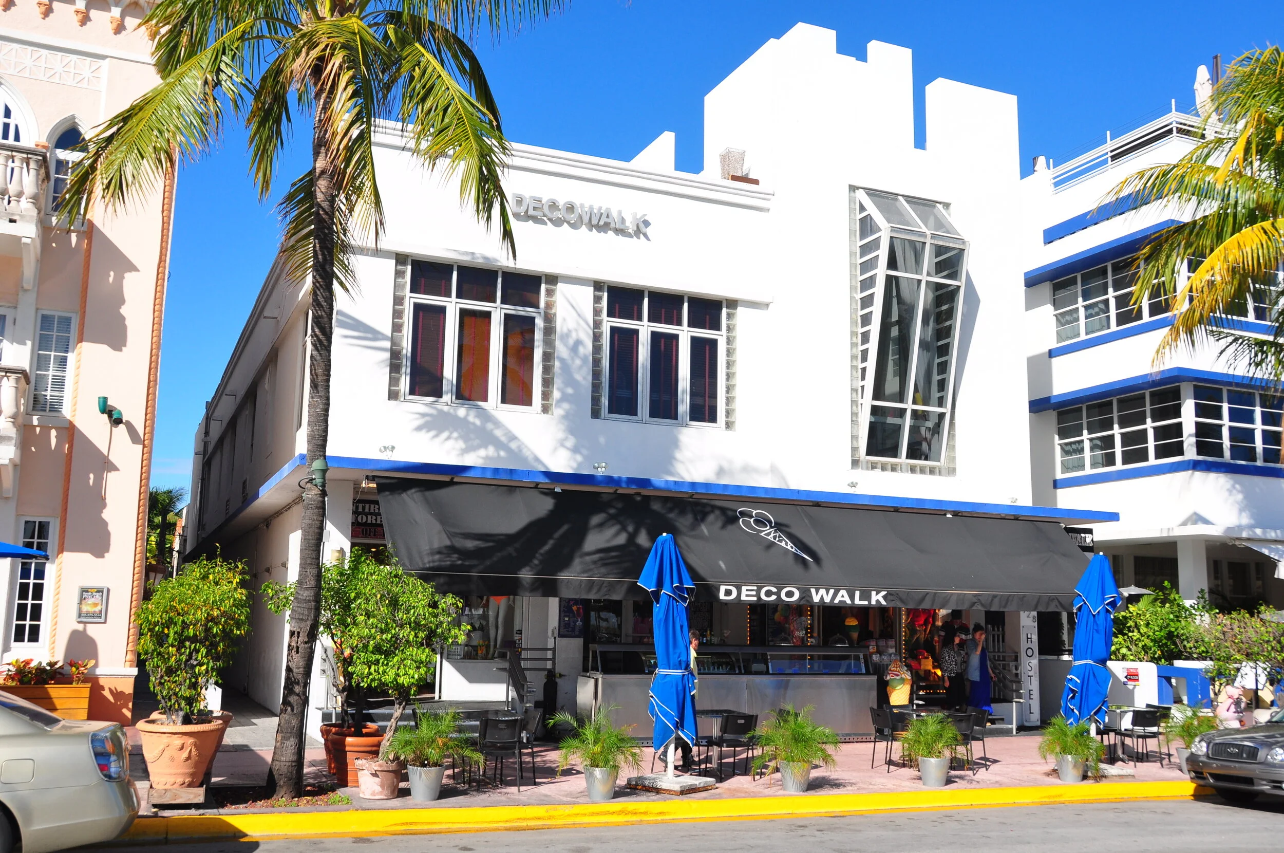 Street view of DecoWalk shopping area with a white modern building, outdoor seating with black tables and blue umbrellas, palm trees, potted plants, and parked cars.