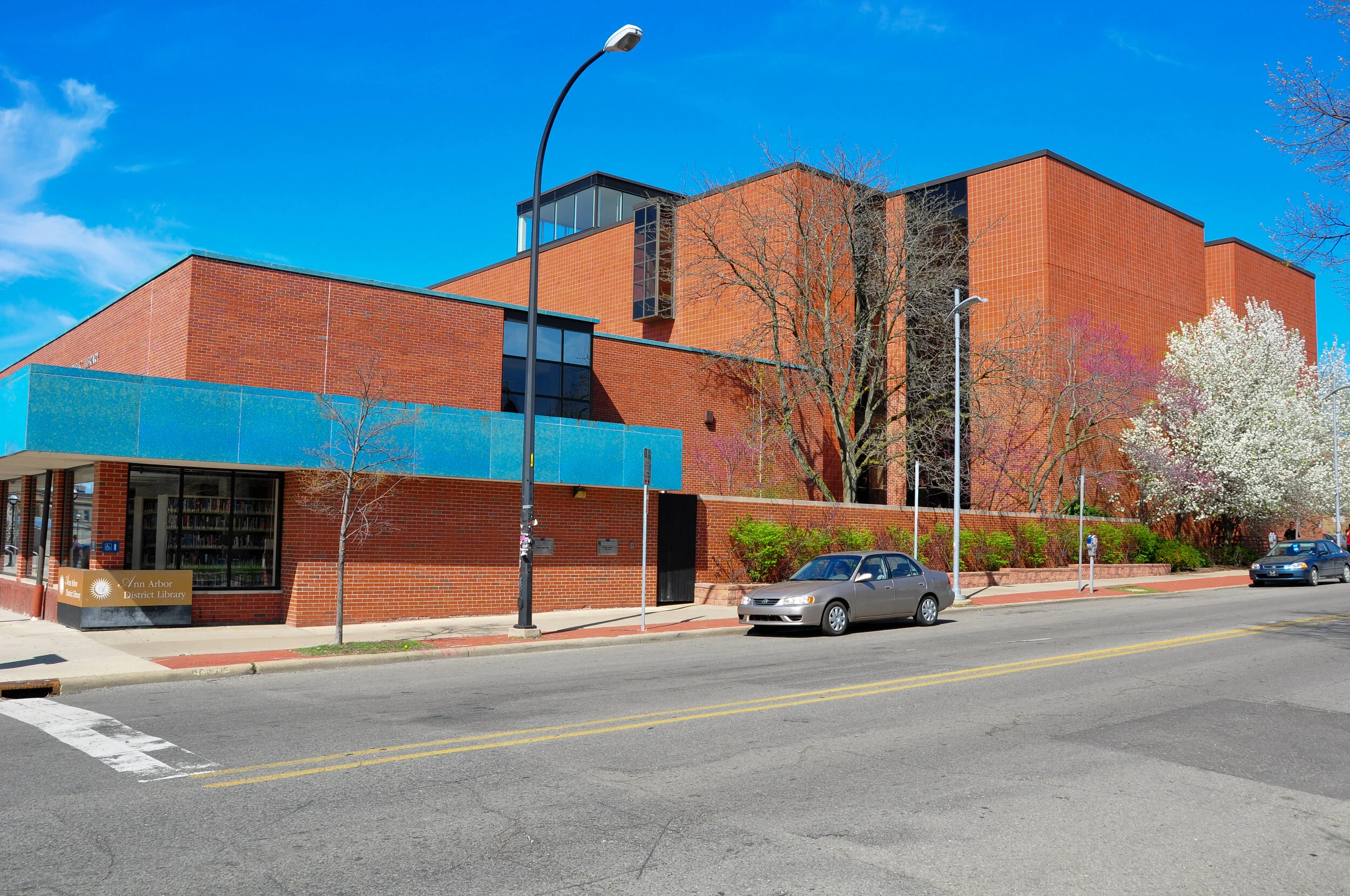 Exterior view of Ann Arbor District Library building with red brick walls, blue accents, trees, and parked cars under a bright blue sky.