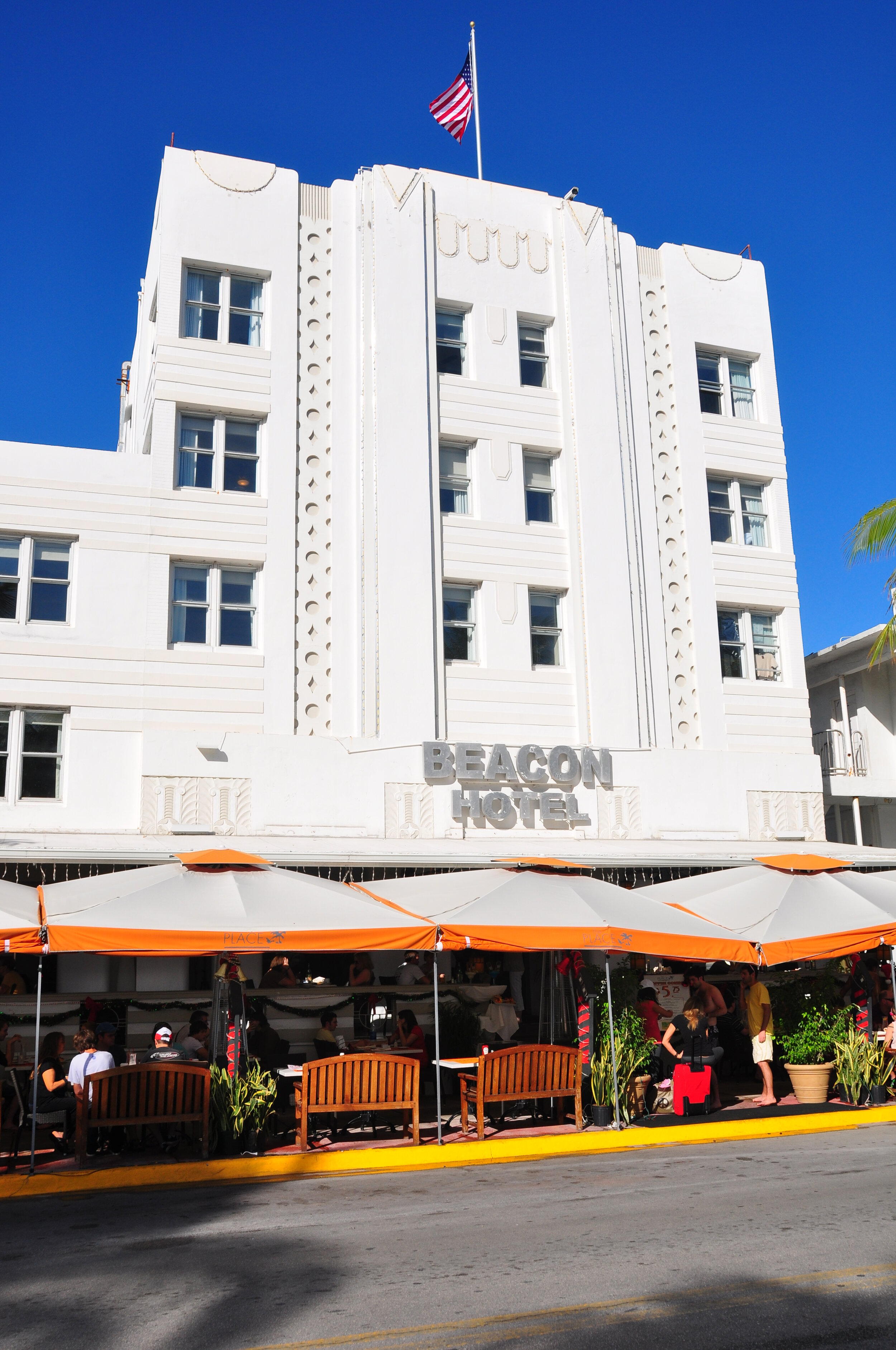 White art-deco hotel building with an American flag on the roof and a sign that reads 'Beacon Hotel'. Outdoor seating area with orange and white umbrellas and people sitting and standing outside.