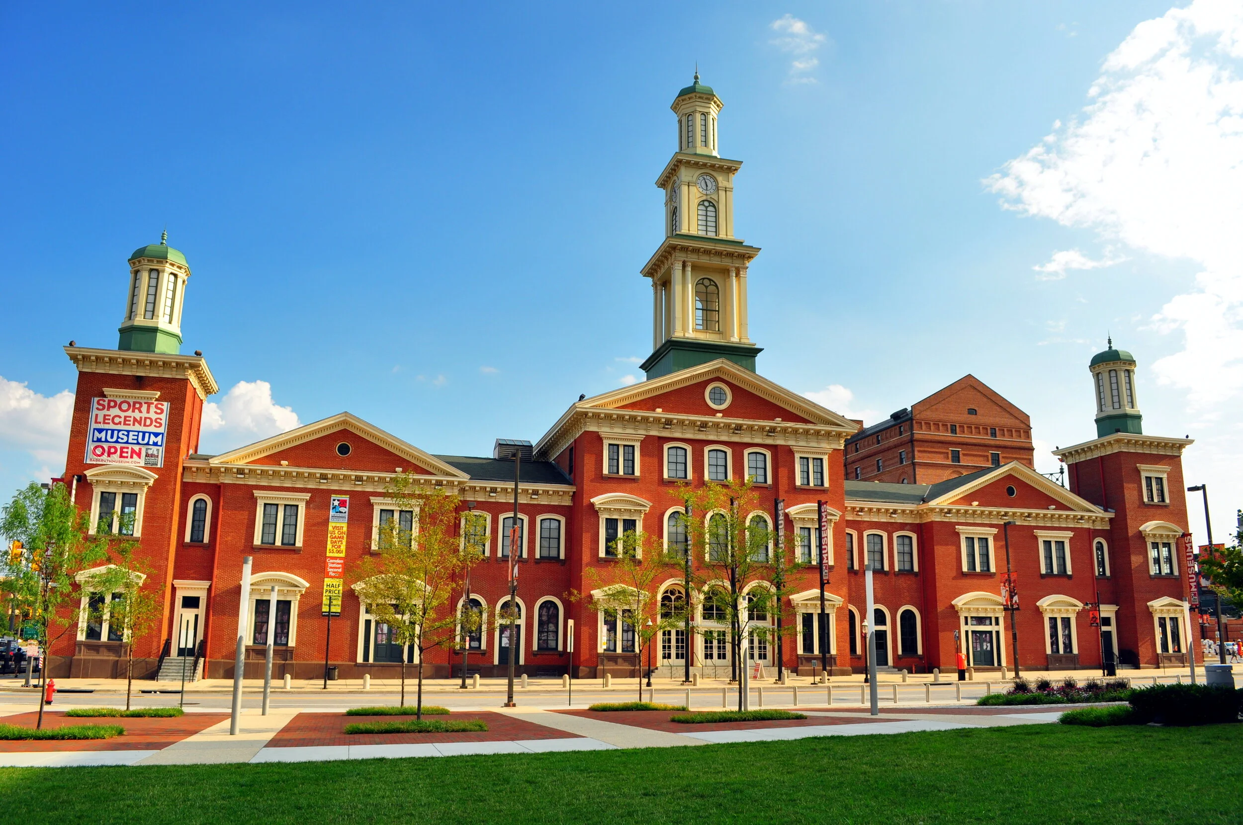 A historic red brick building with a central clock tower and green-domed towers on each side, surrounded by a landscaped park with trees and sidewalks.