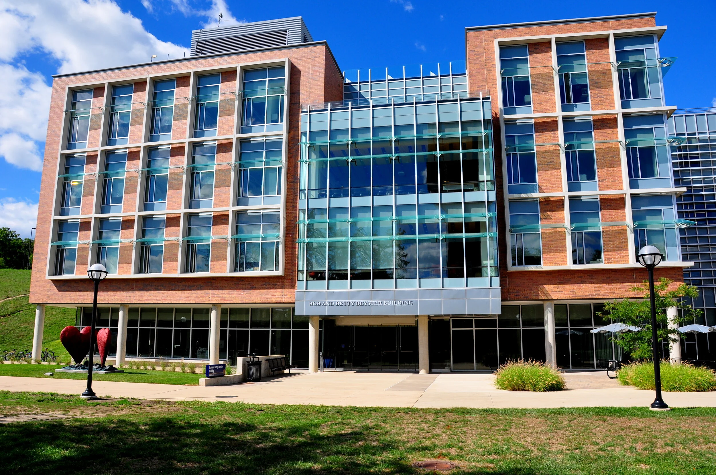 Modern multi-story building with brick and glass facade, labeled 'Bob and Betty Beyster Building', with sculptures of two red hearts on the lawn, lamps, benches, and greenery outside against a partly cloudy sky.