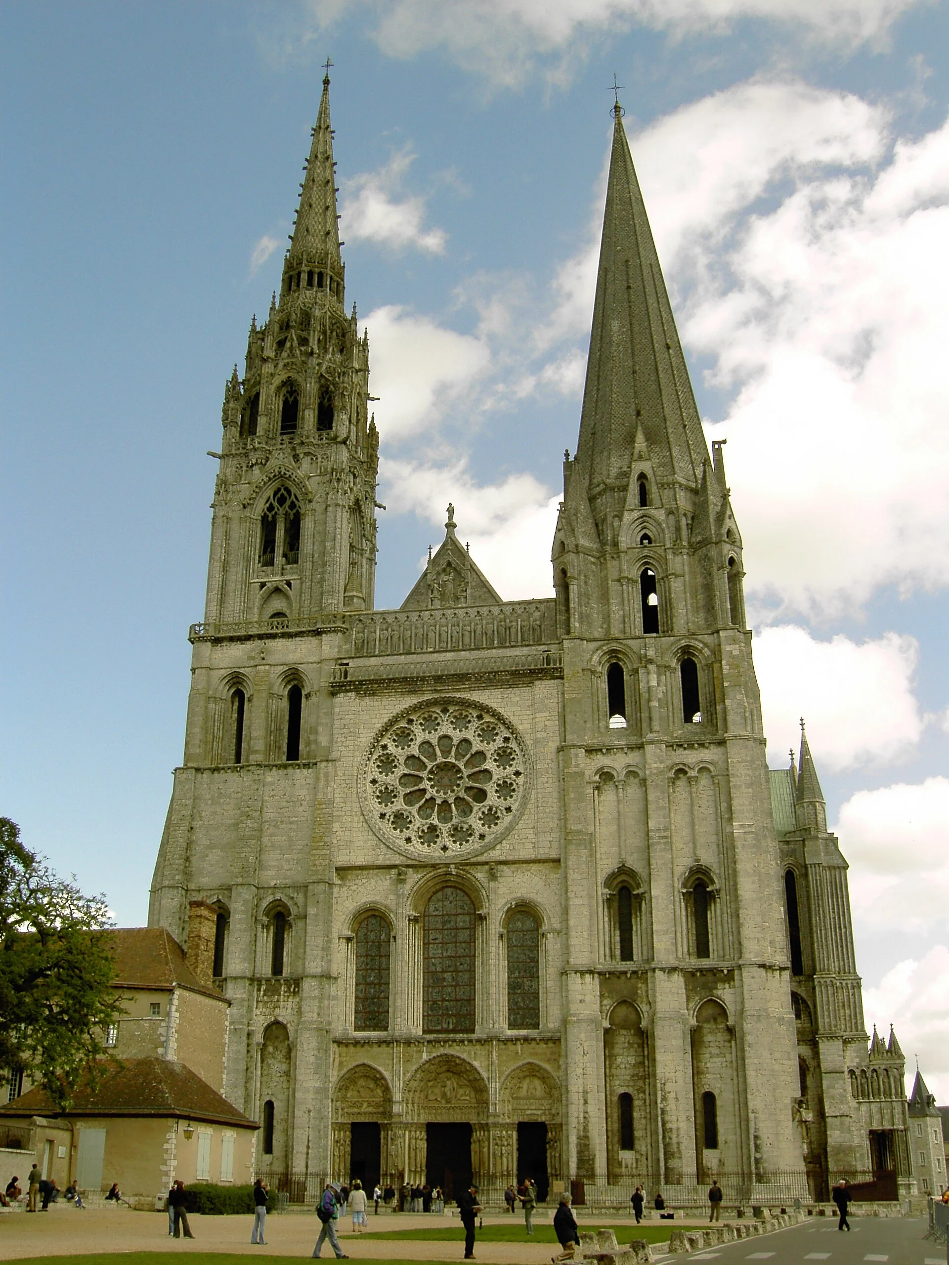 Gothic cathedral with two tall spires and a large rose window, people walking in front of it, blue sky with clouds.