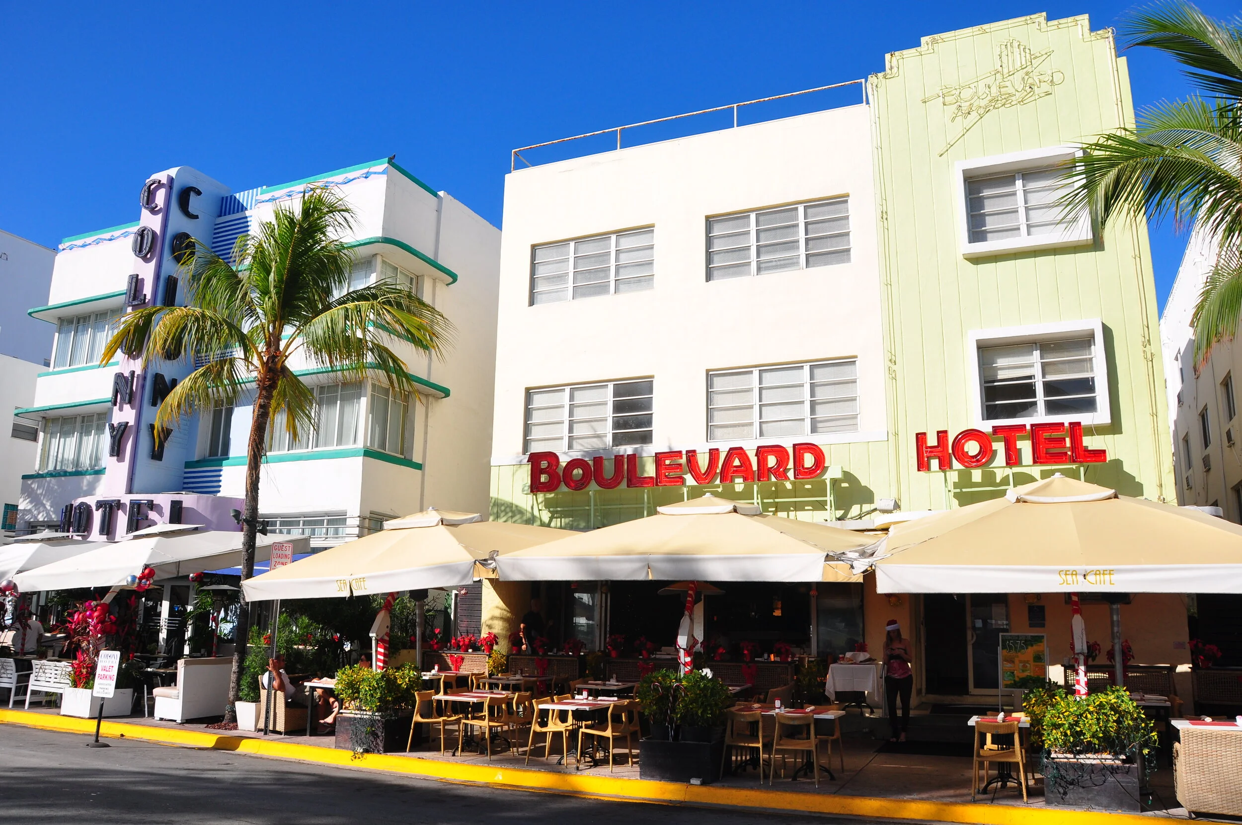 Colorful vintage hotel with neon signs reading 'Boulevard Hotel' and 'Cosmopolitan Hotel', outdoor patio with tables and umbrellas, palm trees, sunny weather, blue sky.