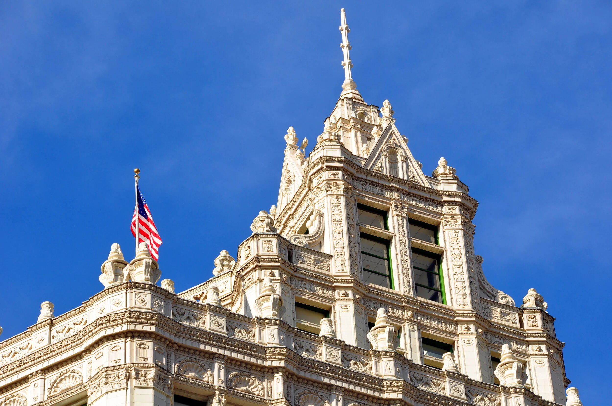 Close-up of an ornate, historic building with decorative carvings, tall windows, and a pointed spire against a clear blue sky; an American flag flying on the roof.