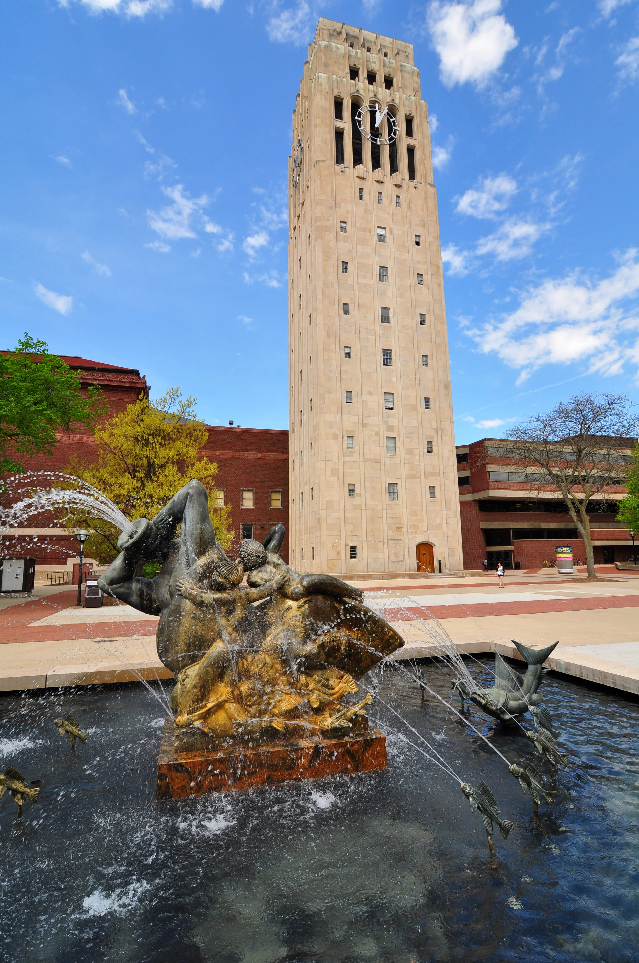 A tall clock tower with a statue fountain in front, featuring a sculpture of children playing, water splashing, and fish sculptures in a plaza with trees and a red brick building.