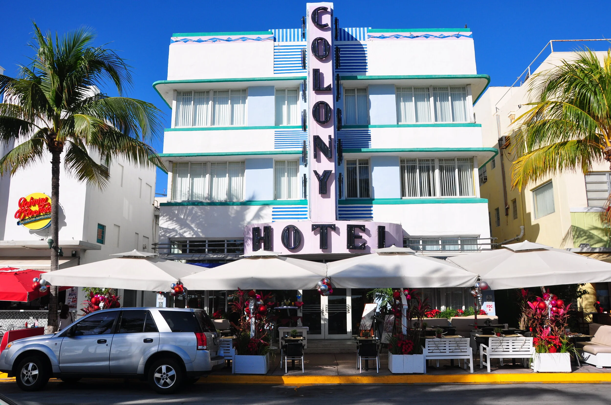 Exterior of a vintage hotel with a pink and purple sign reading 'ColonY Hotel.' The building is white with turquoise accents and large windows. The hotel has outdoor seating with white umbrellas, red and pink decorations, and a silver SUV parked in f
