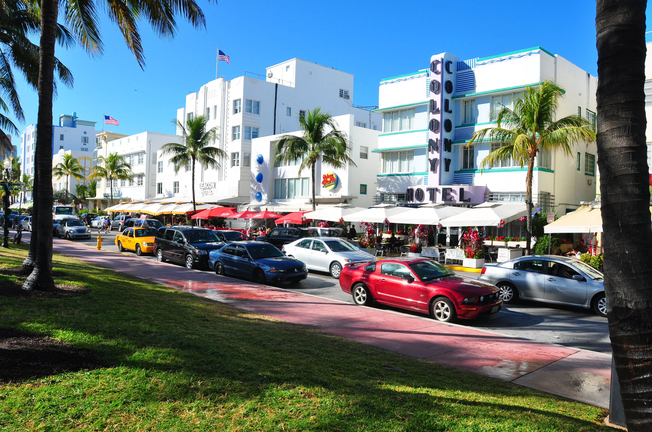Sunny day in a city street with palm trees, parked cars, and a white hotel building with a sign that reads 'Colony Hotel.' Outdoor seating with umbrellas and flowering plants in front of the hotel.