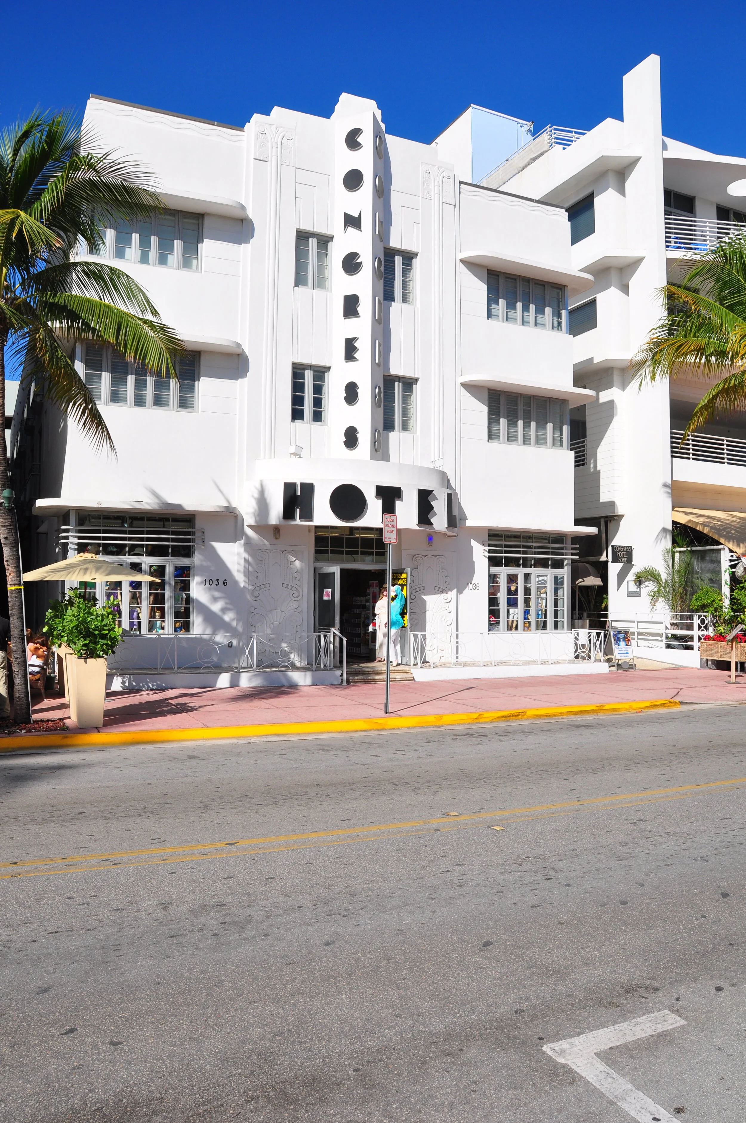 White Art Deco style hotel building with vertical sign reading 'LORRES HOTEL' in black letters, palm trees on sides, clear blue sky overhead, and a pink sidewalk in front.