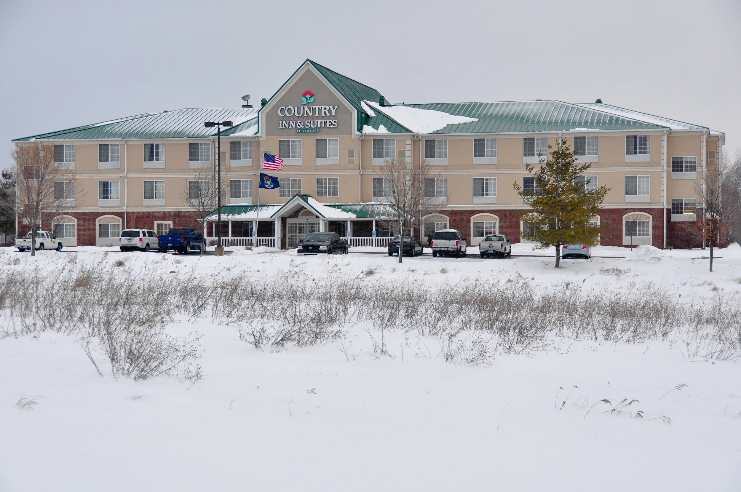 A large hotel called Country Inn & Suites with a green metal roof, beige and brick exterior, and several parked cars in front, surrounded by snow and leafless trees during winter.
