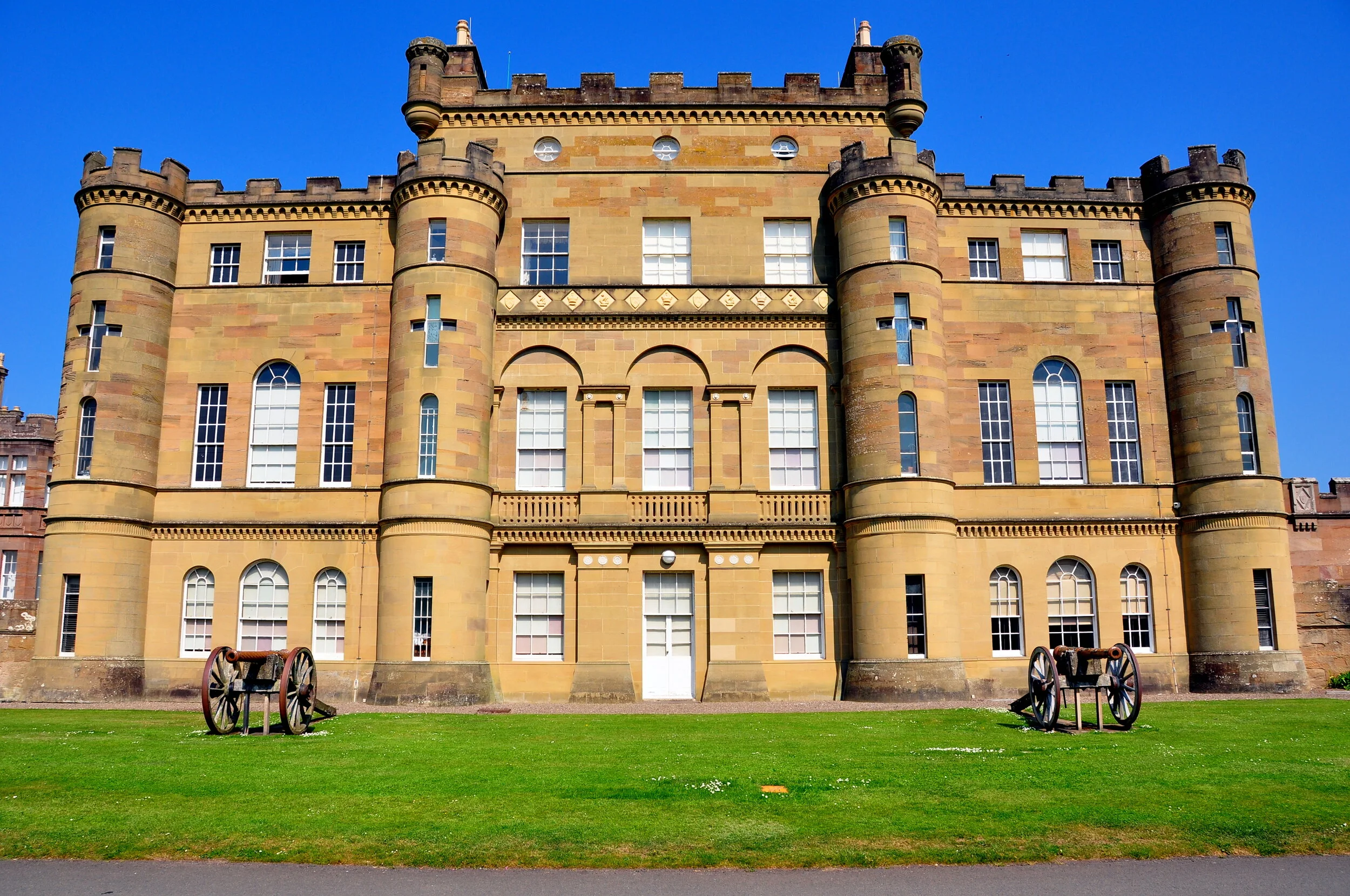 A large historic sandstone castle with towers and turrets under a clear blue sky, surrounded by a lawn with two old cannons in front.