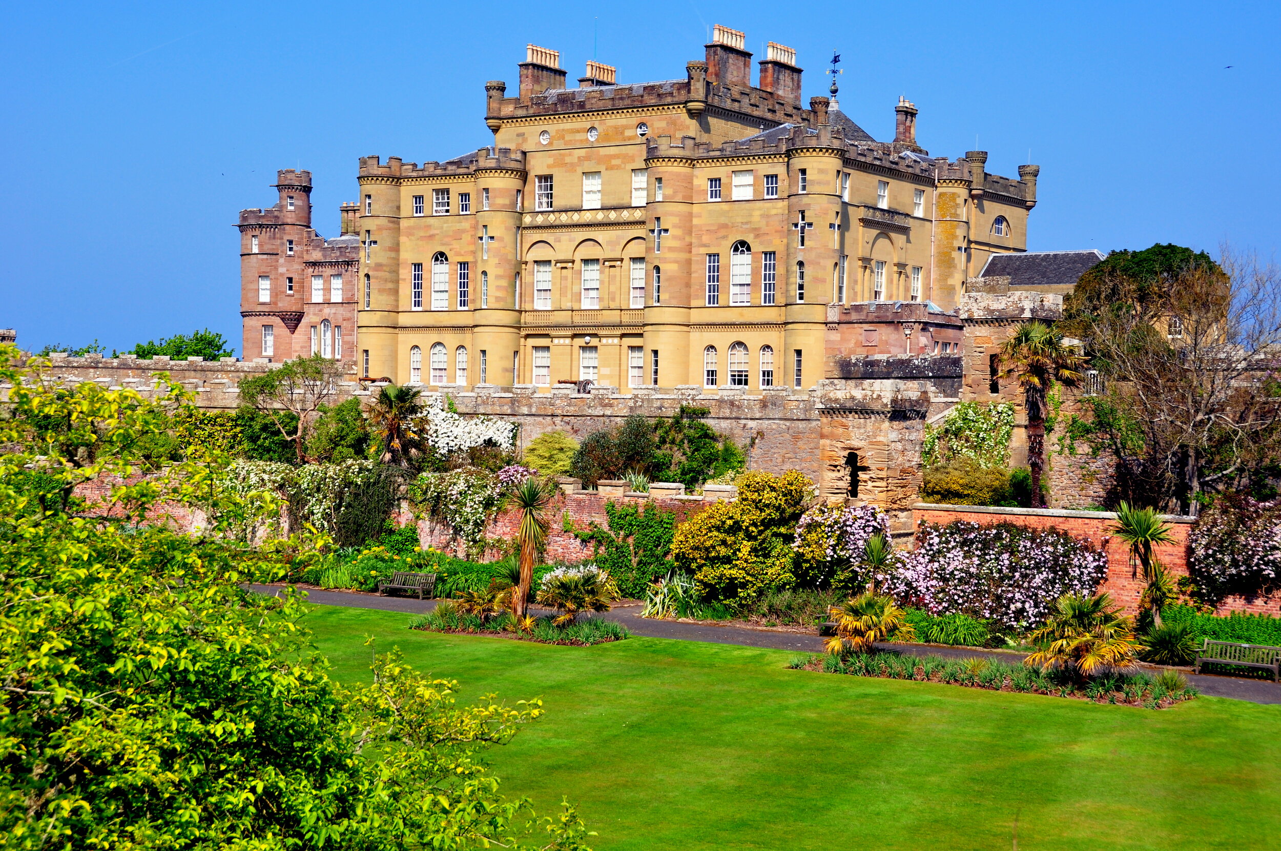 A large historic castle with a stone brick exterior, surrounded by a lush garden with green lawns, palm trees, bushes, and flowering plants, under a clear blue sky.