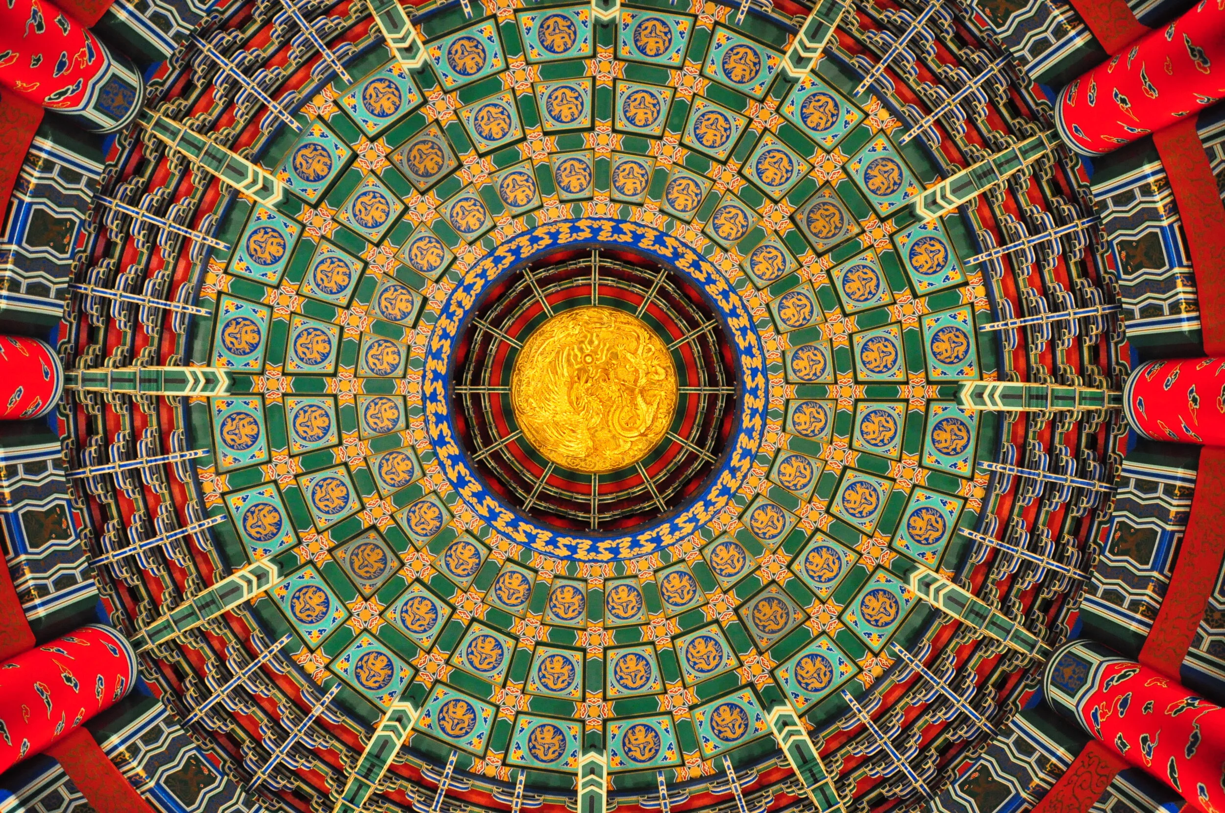 Intricate, colorful ceiling with gold dragon emblem at the center, surrounded by green, blue, red, and gold patterns in an Asian architectural style.