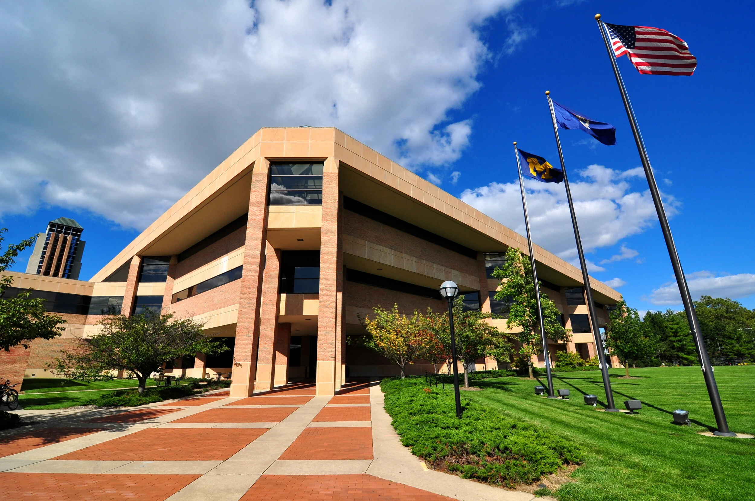 Modern brick building with flags of the United States, a state, and an organization on tall flagpoles outside, surrounded by green trees and a well-maintained lawn under a partly cloudy sky.