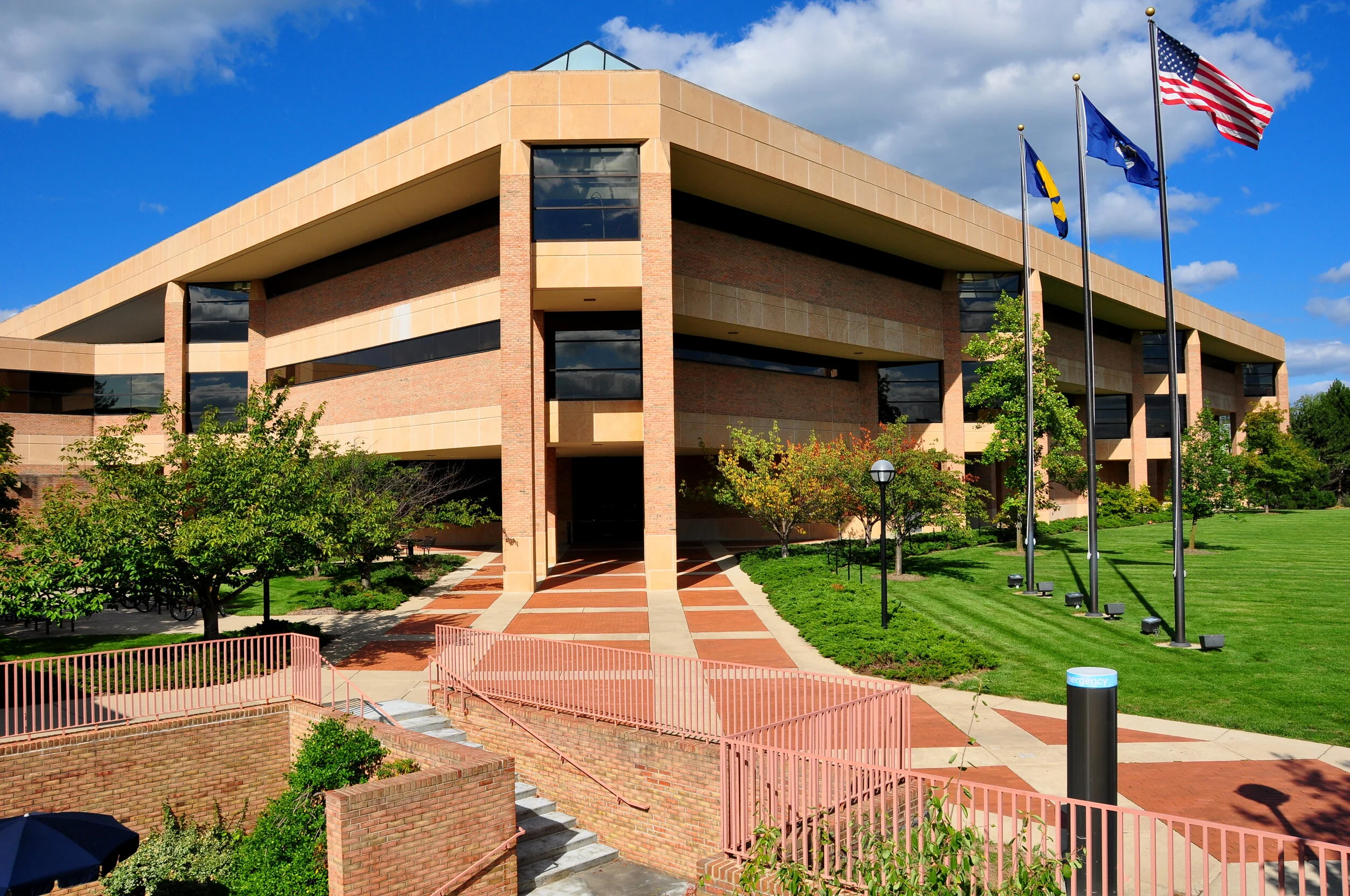 A modern, multi-story building with brick and tan exterior, large glass windows, and surrounded by green trees and grass. There are four flagpoles with flags, including the American flag and others, flying in front.