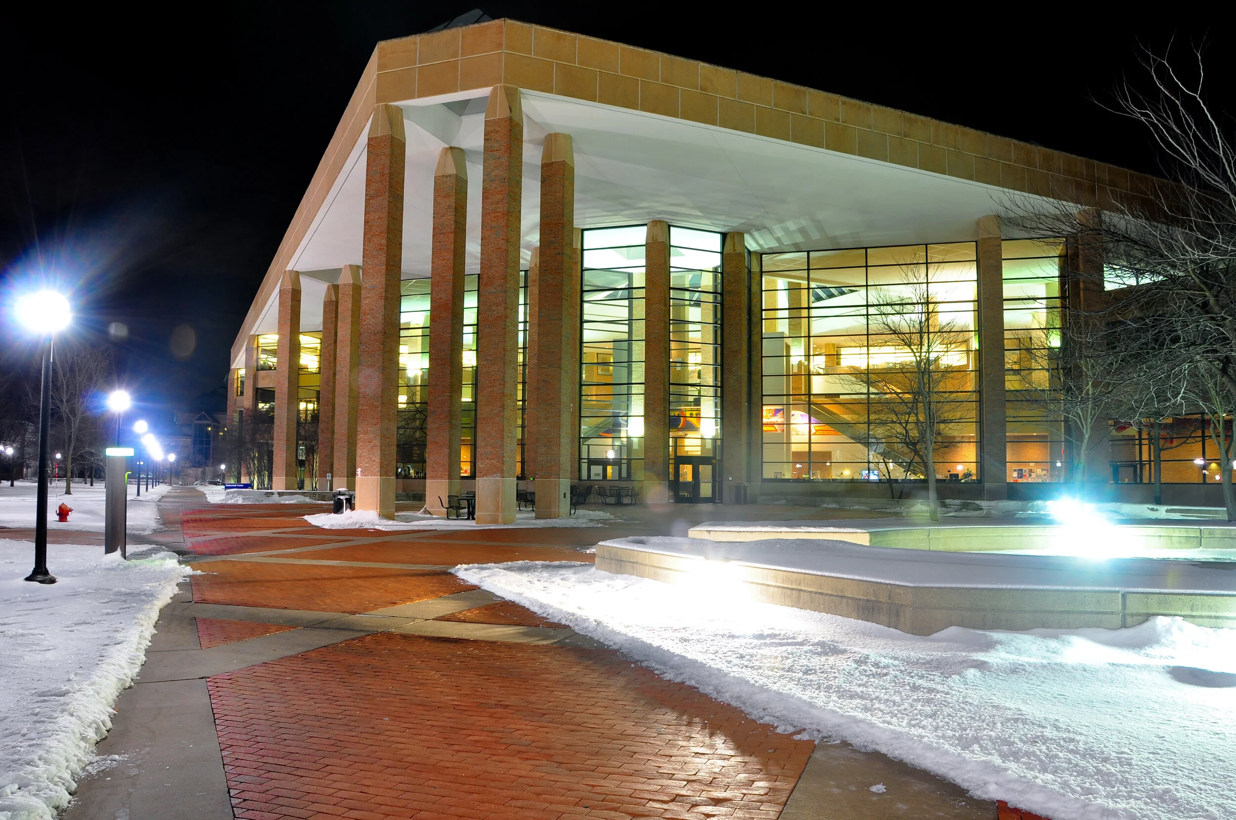 Night view of a modern building with large glass windows, brick accents, and illuminated interior, situated in a winter scene with snow on the ground, street lamps, and leafless trees.