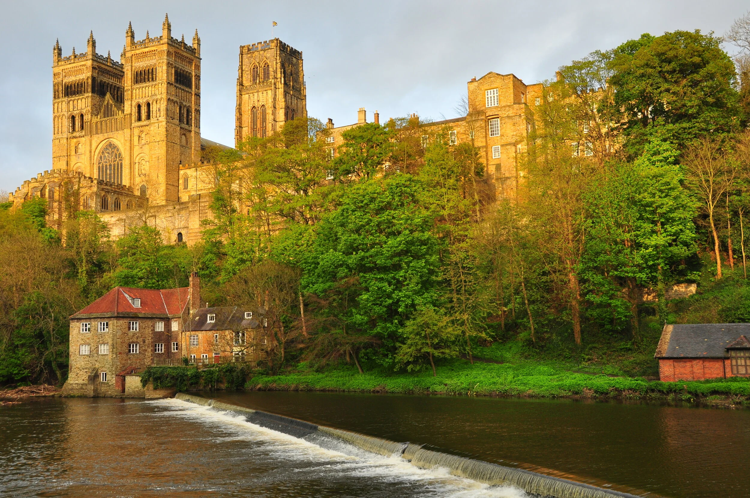 A historic castle situated on a hill surrounded by lush greenery, overlooking a river with a small waterfall and a stone building with a red roof in the foreground, during late afternoon sunlight.