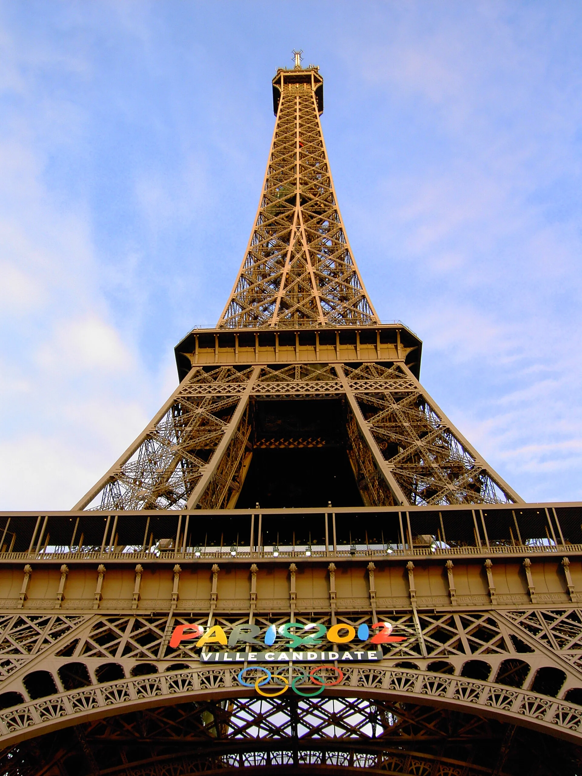 Low-angle view of the Eiffel Tower with a banner reading "Paris 2024 Ville Candidate" and Olympic rings, set against a blue sky.