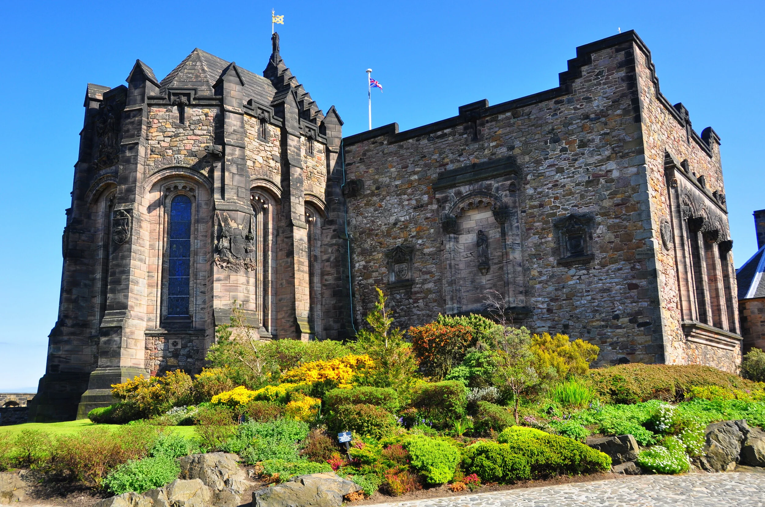 A historic stone castle with tall towers and large stained glass windows, surrounded by colorful manicured garden with bushes and flowers, under a clear blue sky.