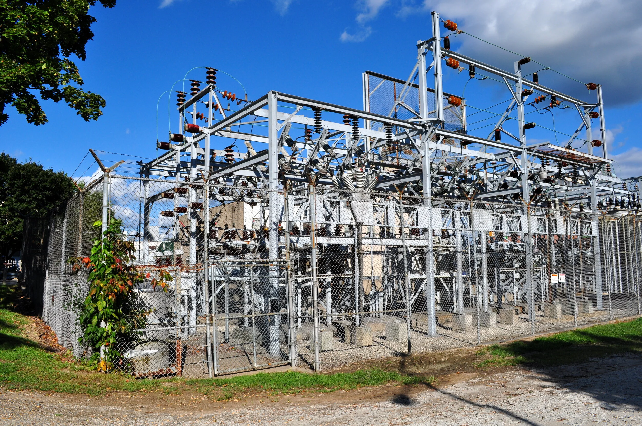 A large electrical substation with metal framework, transformers, insulators, and high-voltage lines, surrounded by a chain-link fence against a partly cloudy sky.