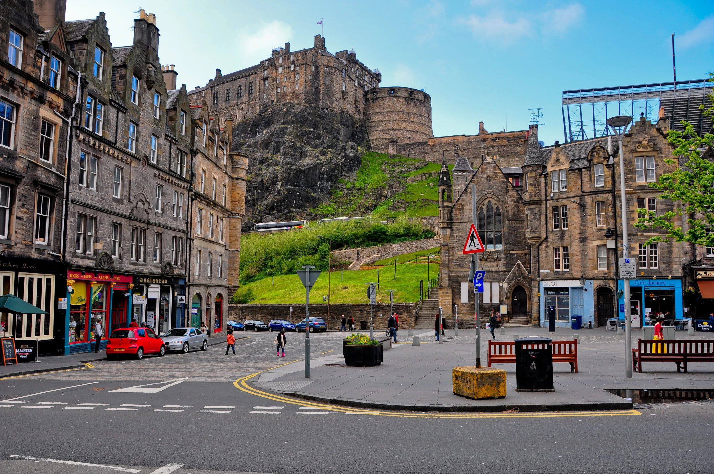 A street scene with multi-story buildings on either side, with a castle atop a hill in the background. People are walking on the sidewalk, and parked cars are visible along the street. There are benches, street signs, and a flower planter in the fore
