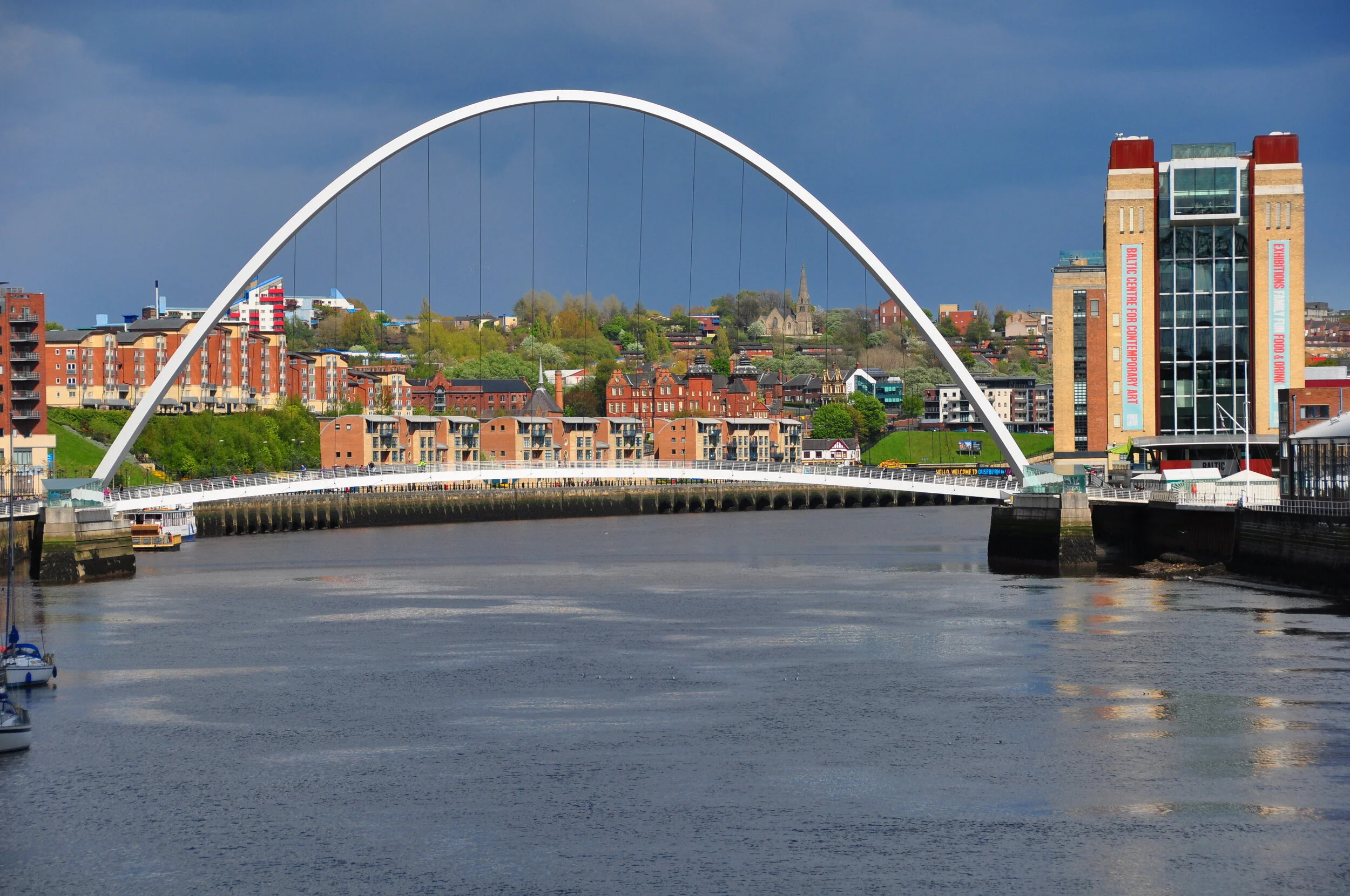 A cityscape featuring a large, white arch bridge over a river, with buildings and greenery on the hillside in the background.