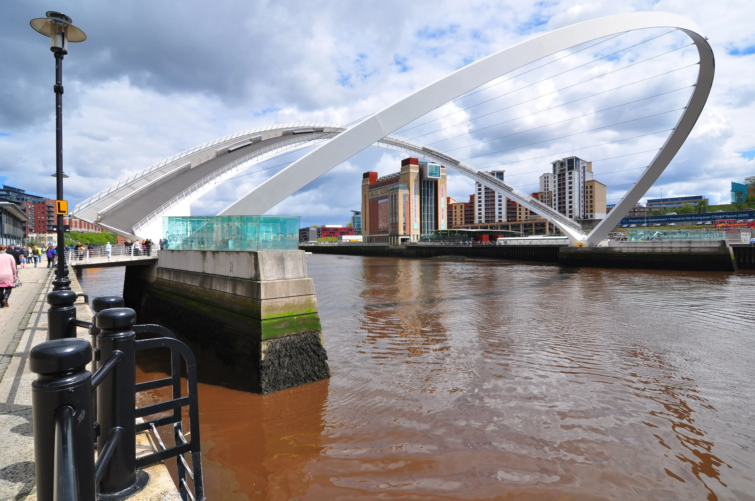 The image shows the Millennium Bridge over the River Tyne in Newcastle, England, with modern buildings in the background and a cloudy sky overhead.