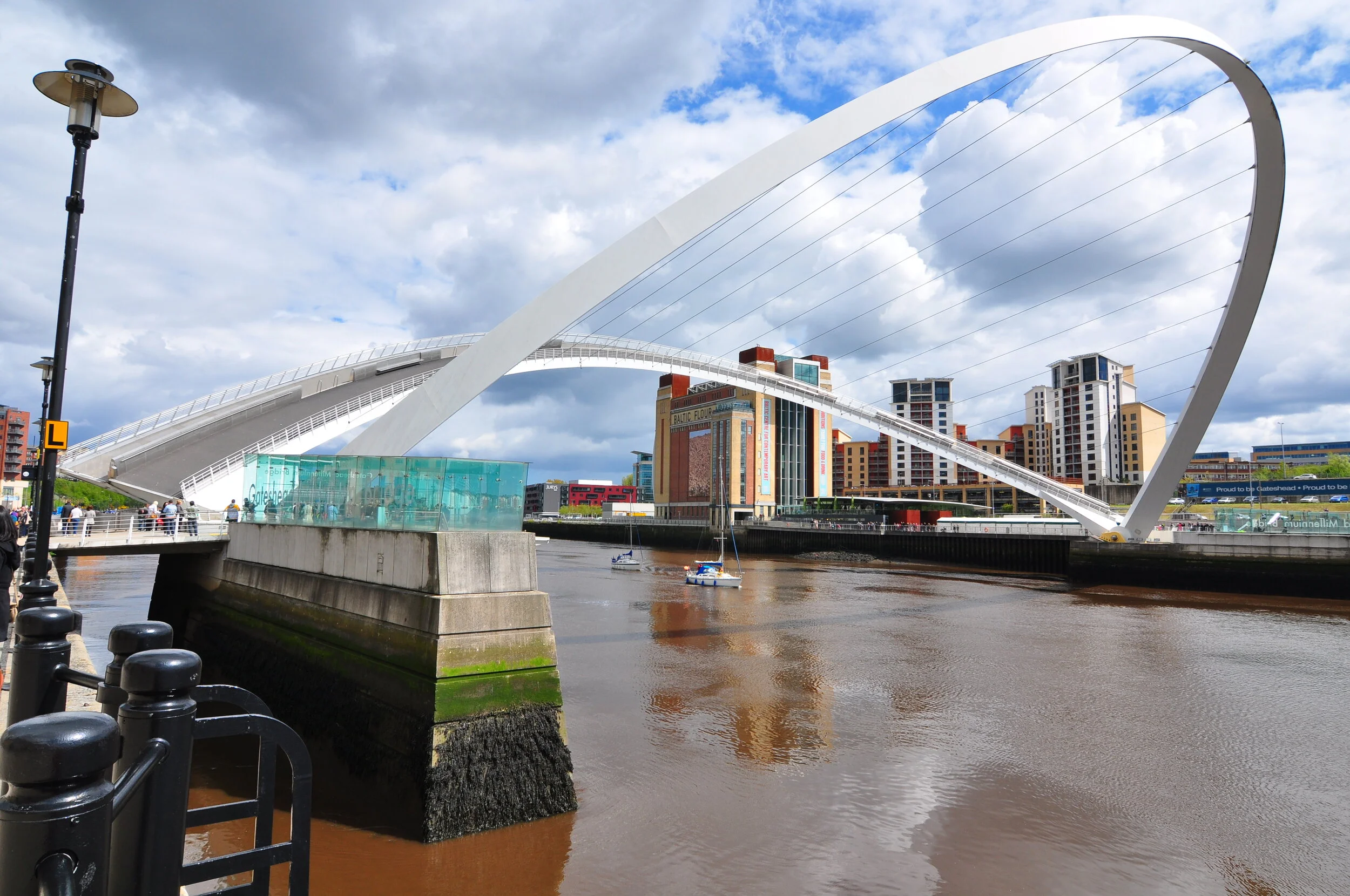 The Millennium Bridge, a modern white suspension pedestrian bridge, spans the River Thames in London with urban buildings in the background under a partly cloudy sky.