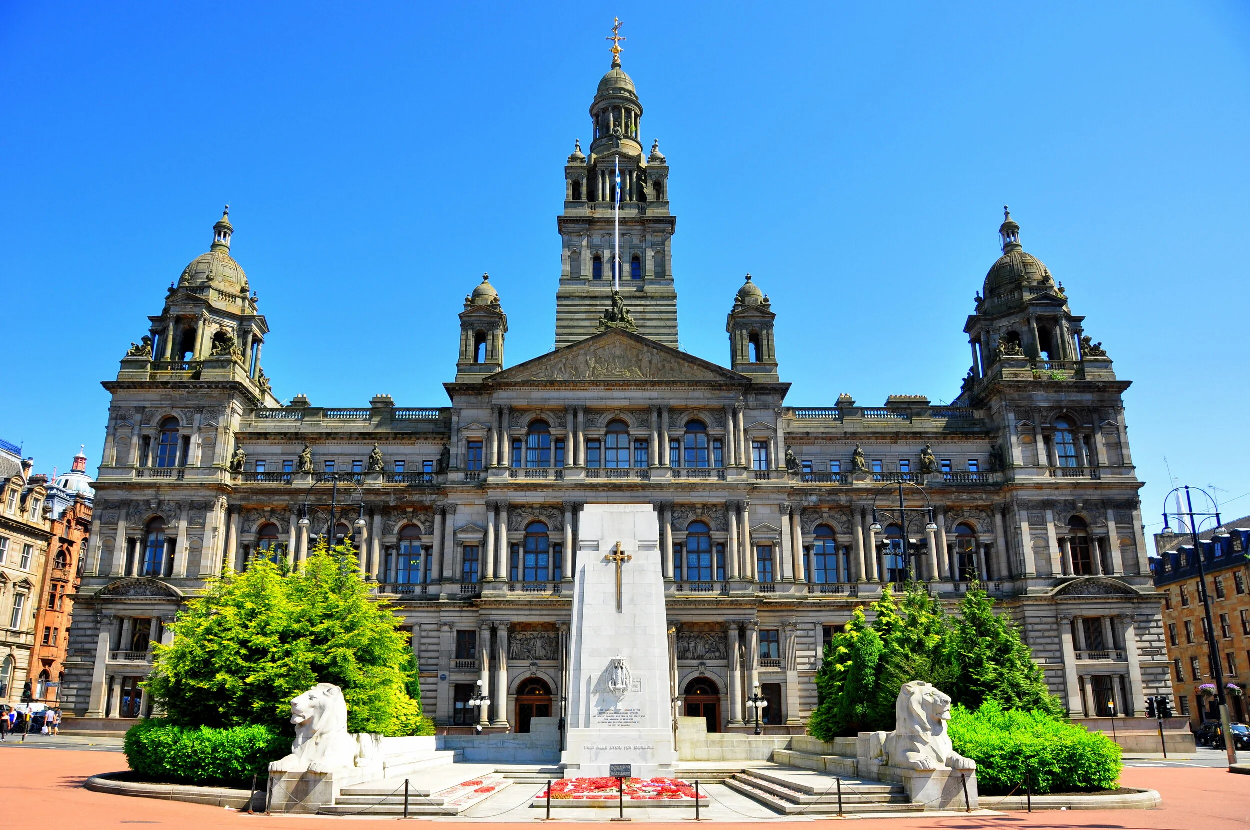 Front view of a historic government building with multiple towers, flags, and a monument with lion statues and flowers in the foreground.