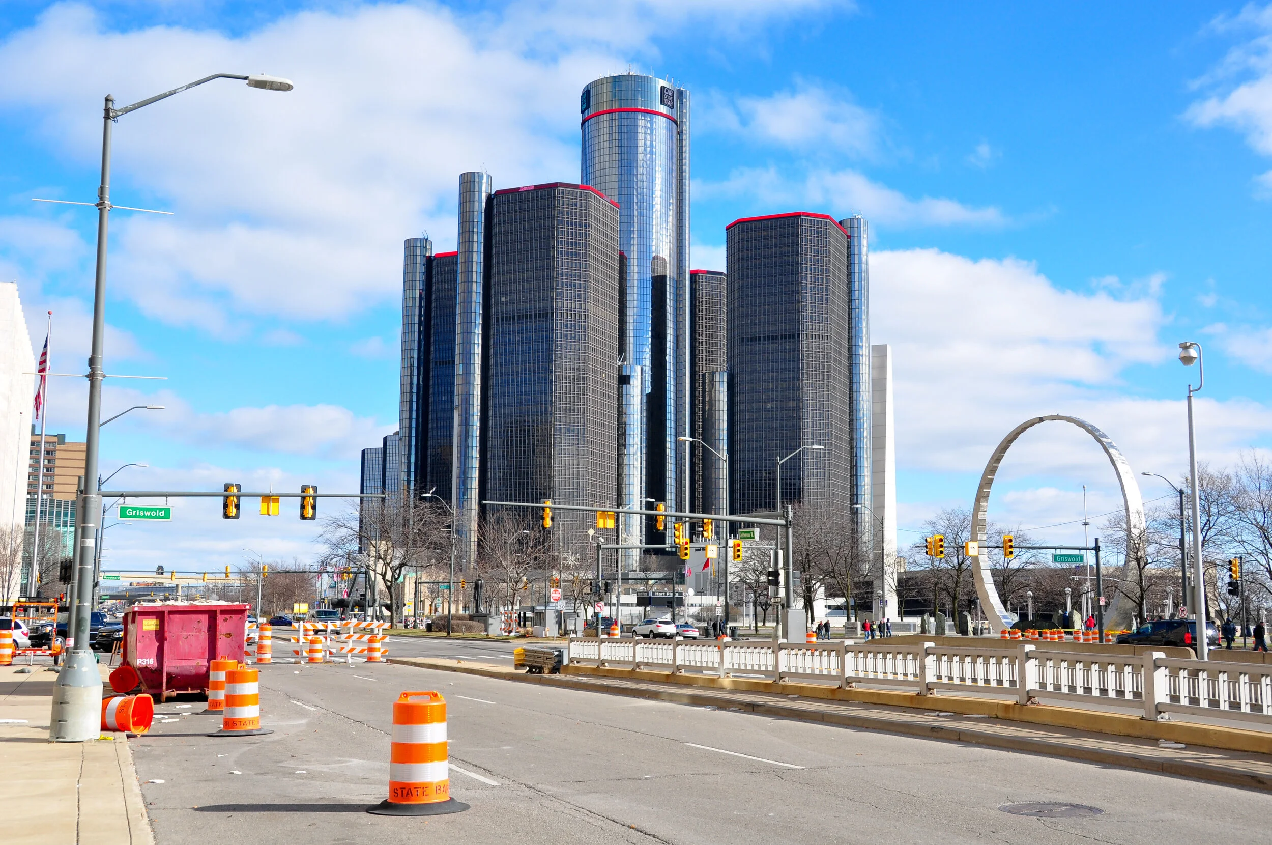 Downtown skyscraper area with a large modern glass building, traffic lights, construction cones, and a decorative circular structure in the background under a partly cloudy sky.