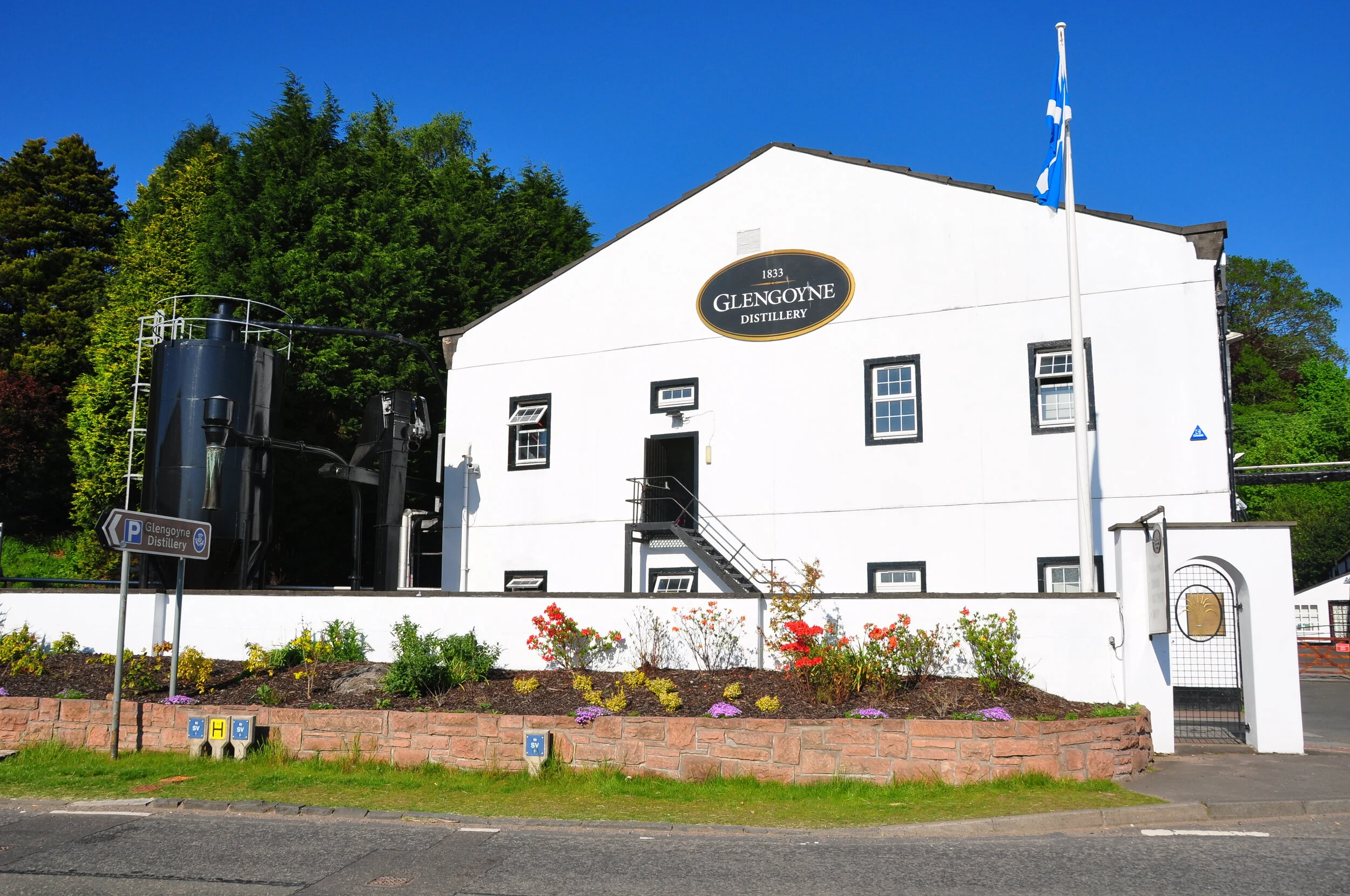 White building labeled Glengoyne Distillery with black trim around windows, a logo sign above the door, a flagpole with a blue and white flag, surrounded by greenery and a flower garden, under a clear blue sky.