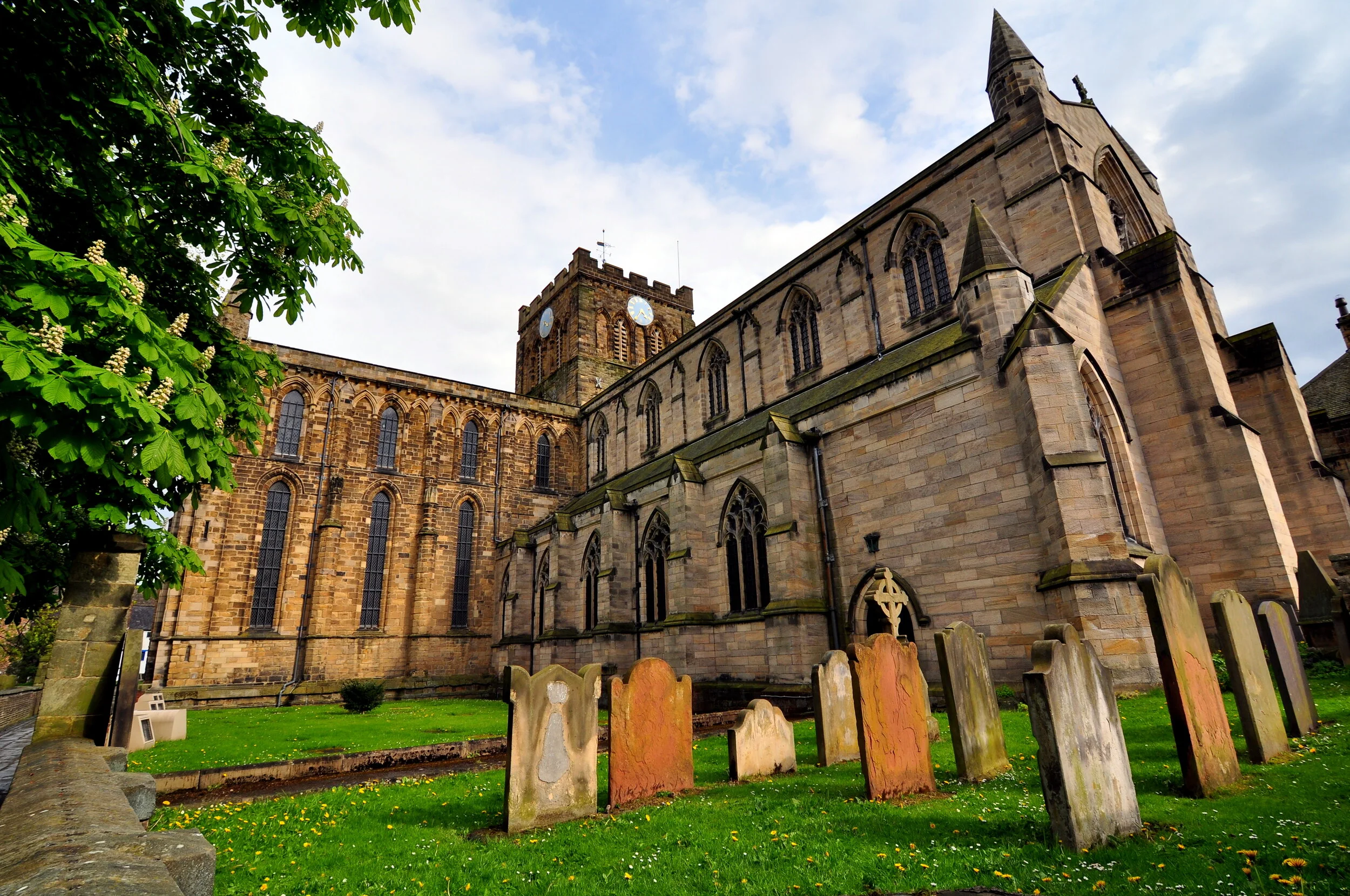 An old stone church with tall arched windows, a clock tower, and a graveyard with weathered headstones in the foreground. Green grass and trees are also visible.