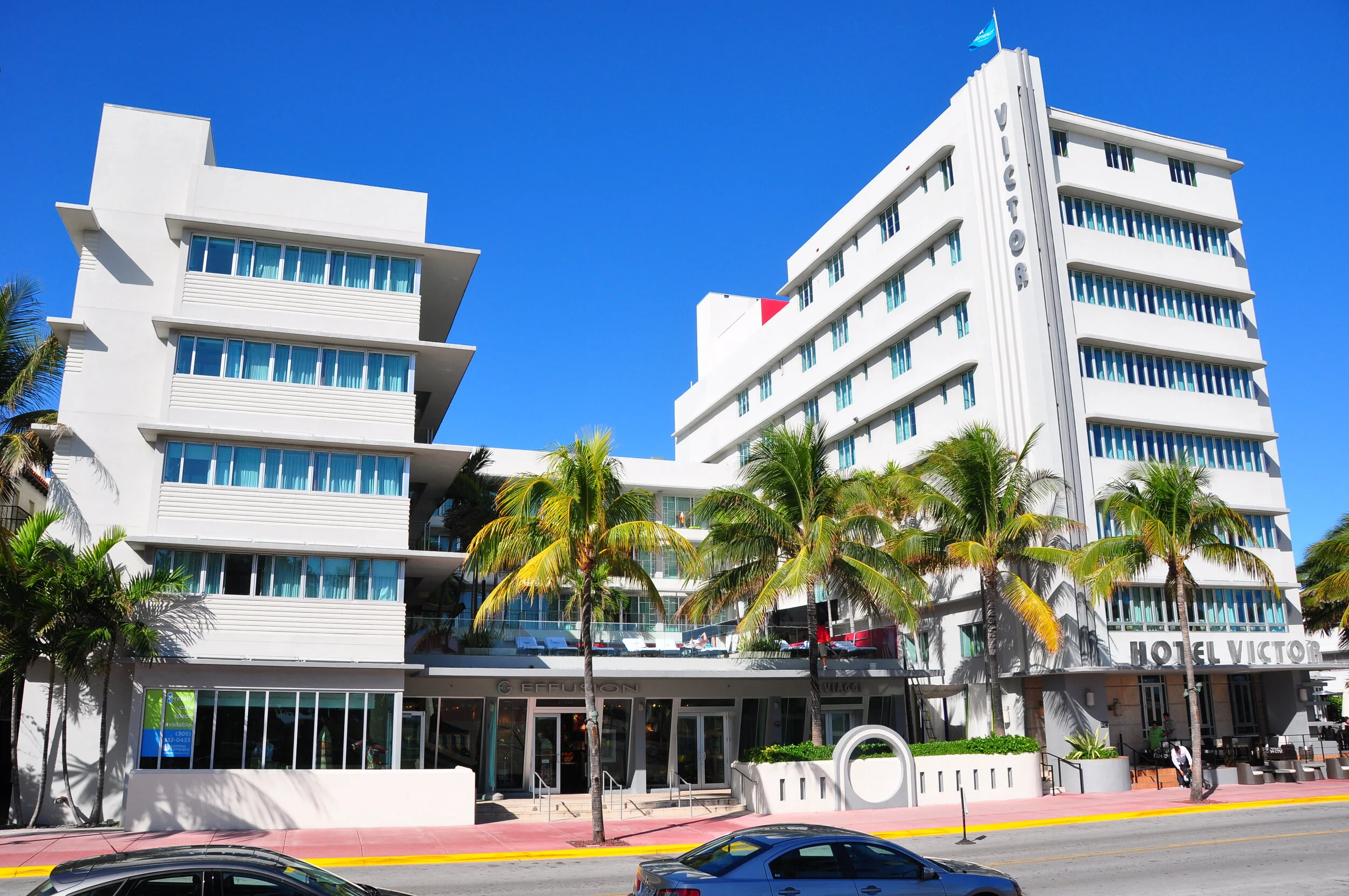 White hotel building with multiple floors and blue windows, surrounded by palm trees, under a clear blue sky.