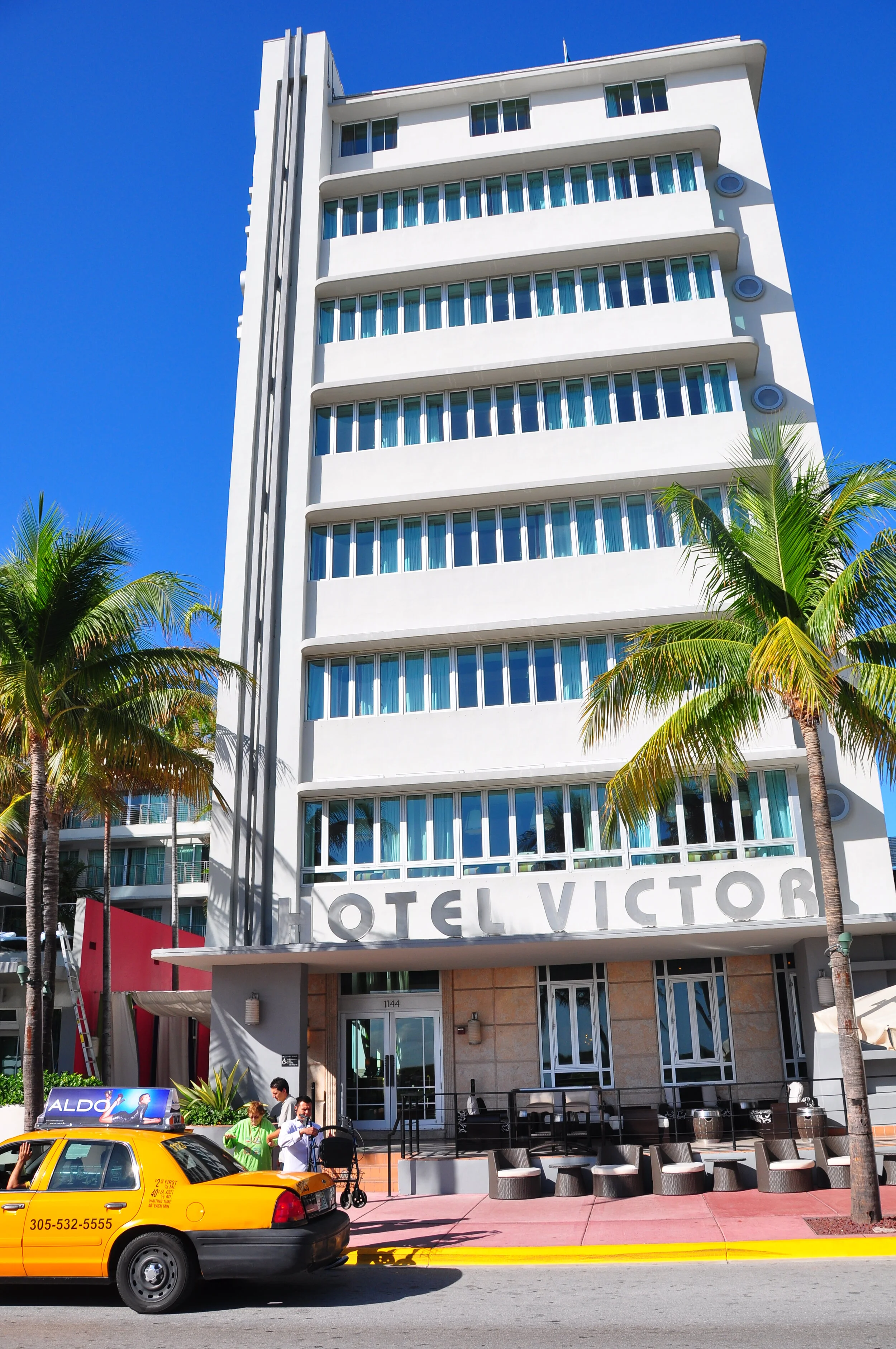 Front view of Hotel Victor, a tall white building with multiple floors, large windows, and palm trees in front. A yellow taxi and people are visible on the sidewalk.