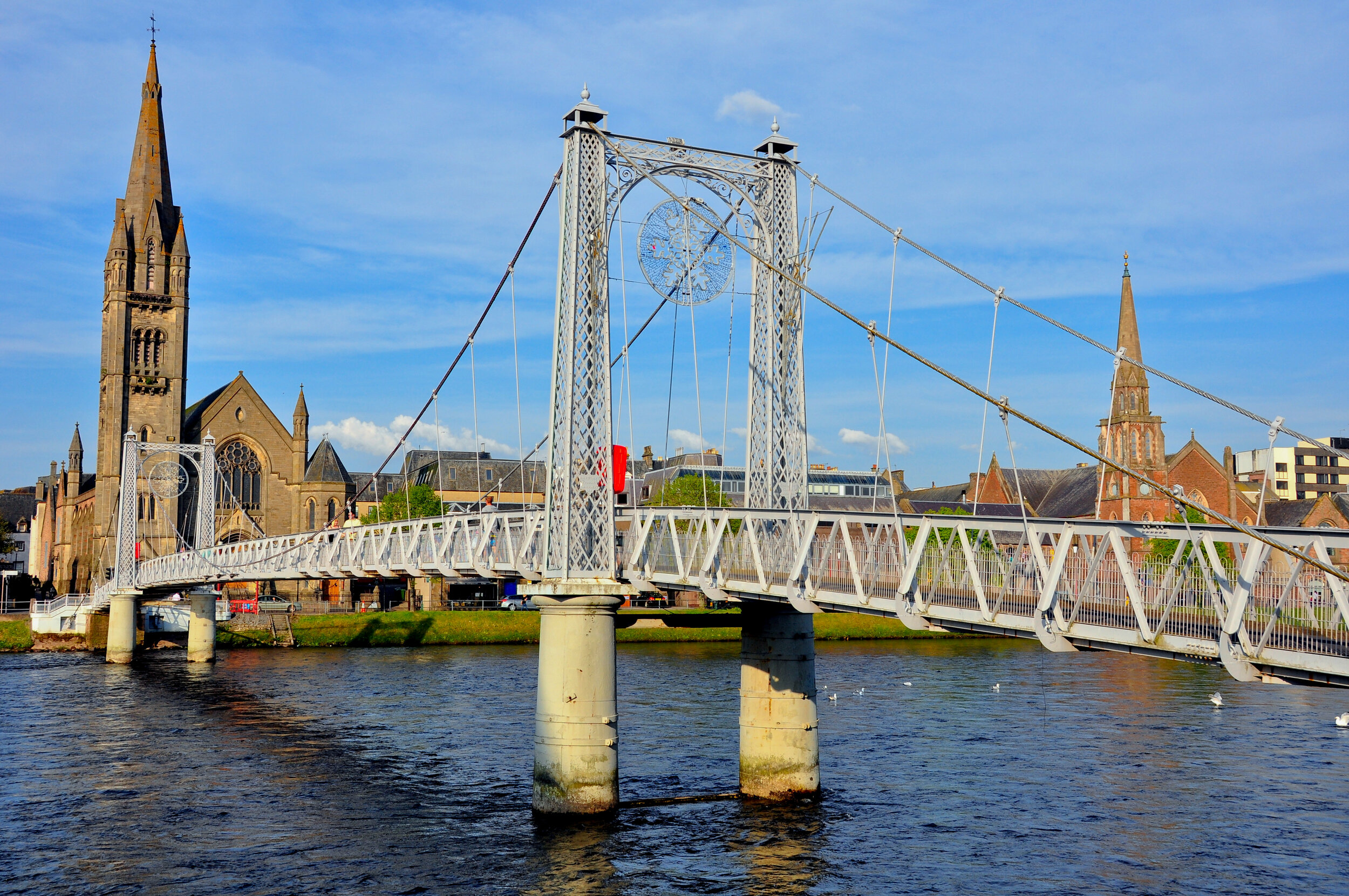 A white suspension footbridge crossing a river with a historic cityscape and church spires in the background.