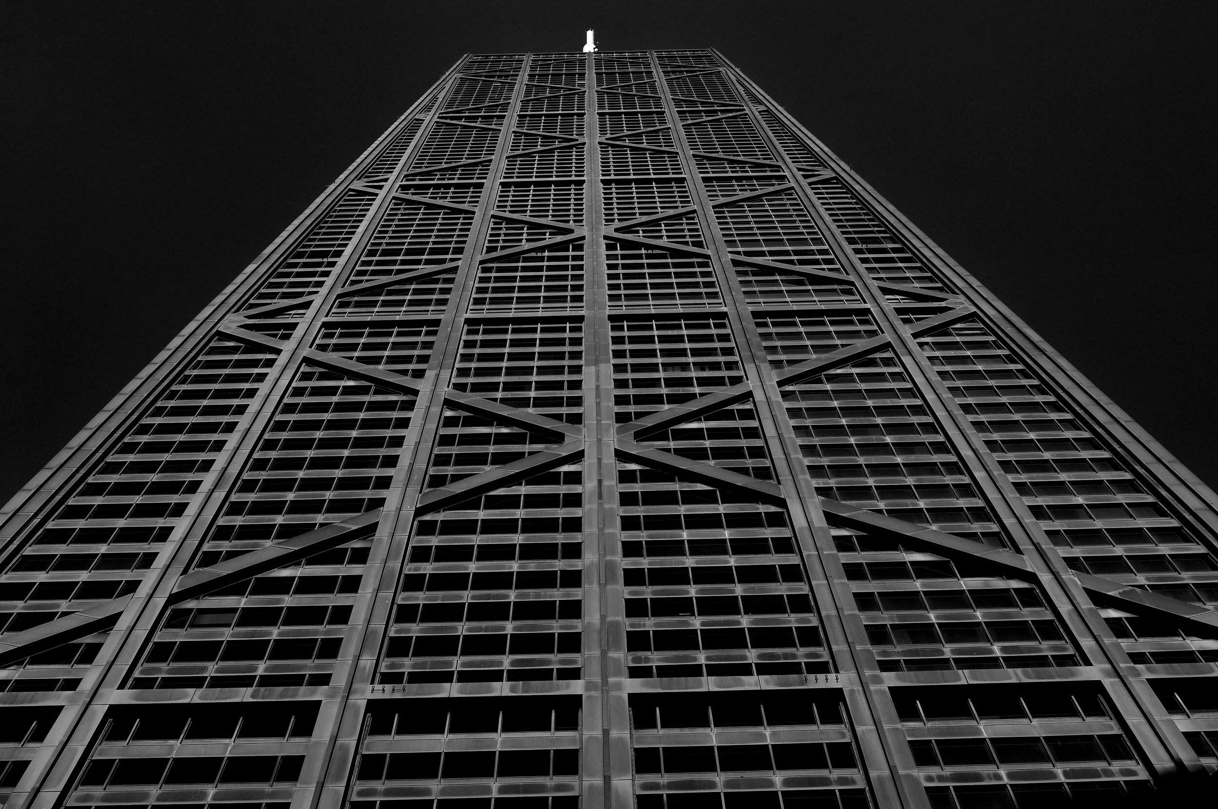A black and white photograph of a tall skyscraper taken from the ground looking upwards, showing its grid-like facade and structural beams.