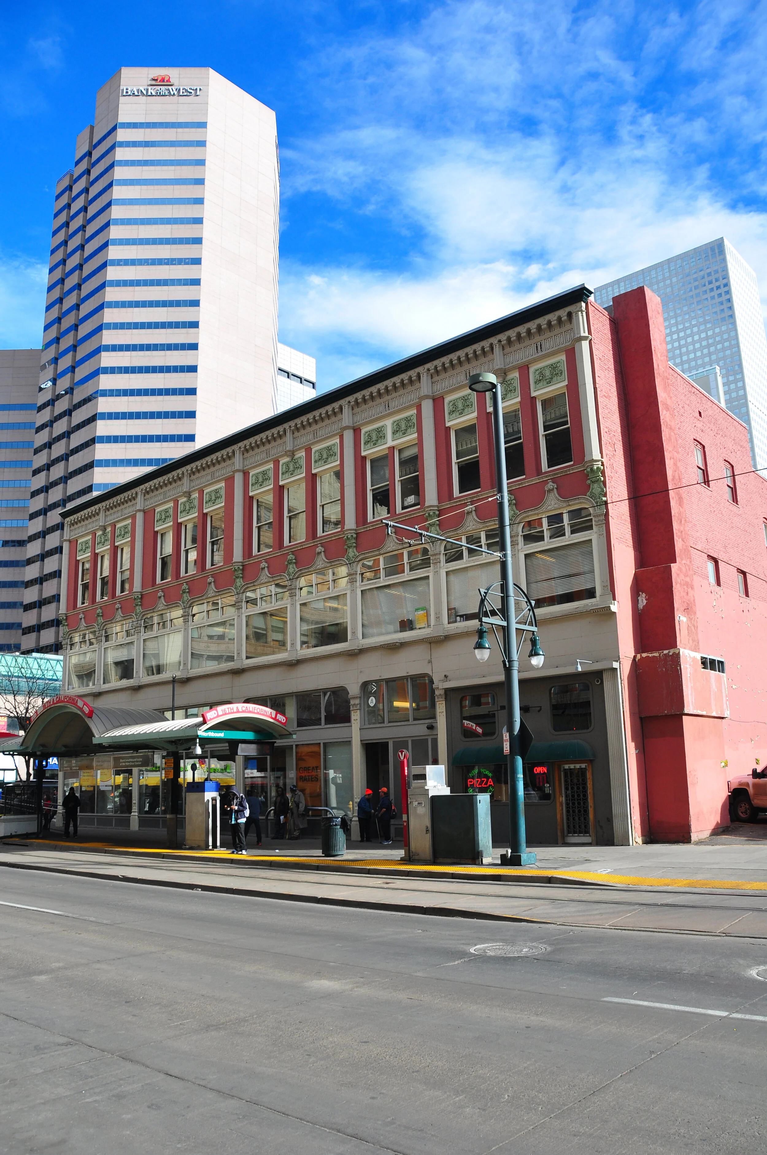 A city street scene featuring a historic pink building with decorative green and white accents, modern skyscrapers in the background, and a bus stop with people waiting.