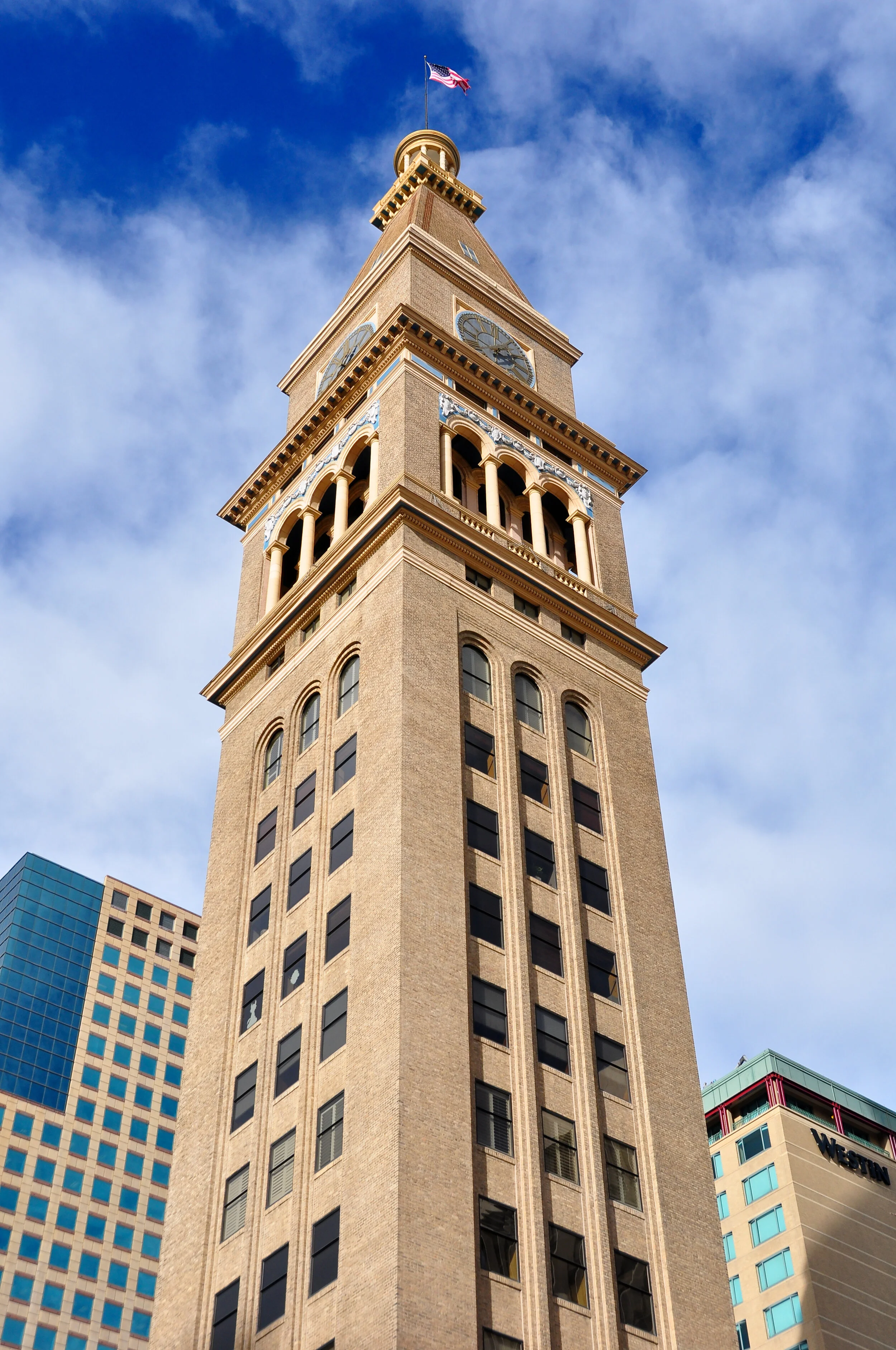 Looking up at a tall historic clock tower with a flagpole and flag, surrounded by modern high-rise buildings, under a partly cloudy blue sky.