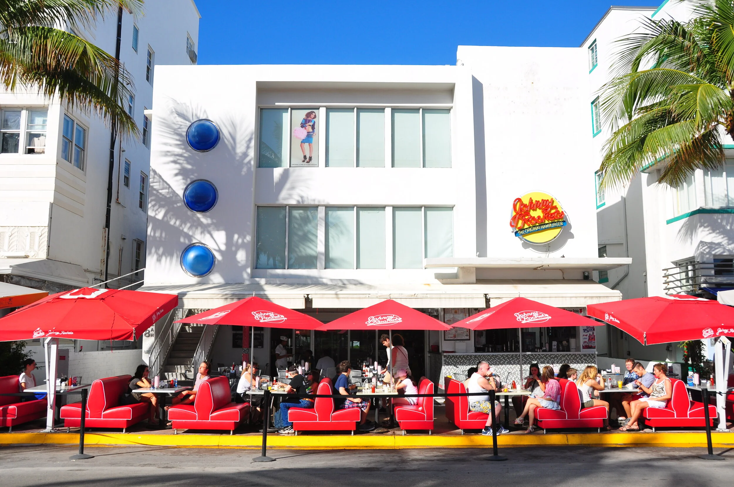 Outdoor dining scene in front of a white building with a "Johnny Rockets" sign, red booths, umbrellas, and diners under sunny skies with palm trees.